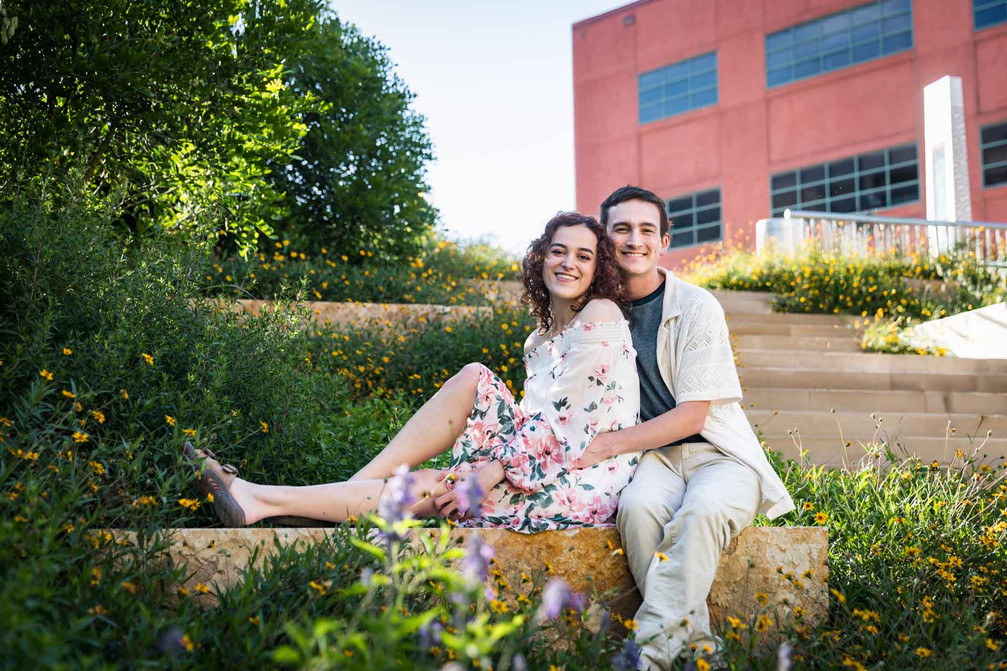 Downtown San Antonio engagement photos of couple seated on stone bench in front of green bushes in San Pedro Creek Park