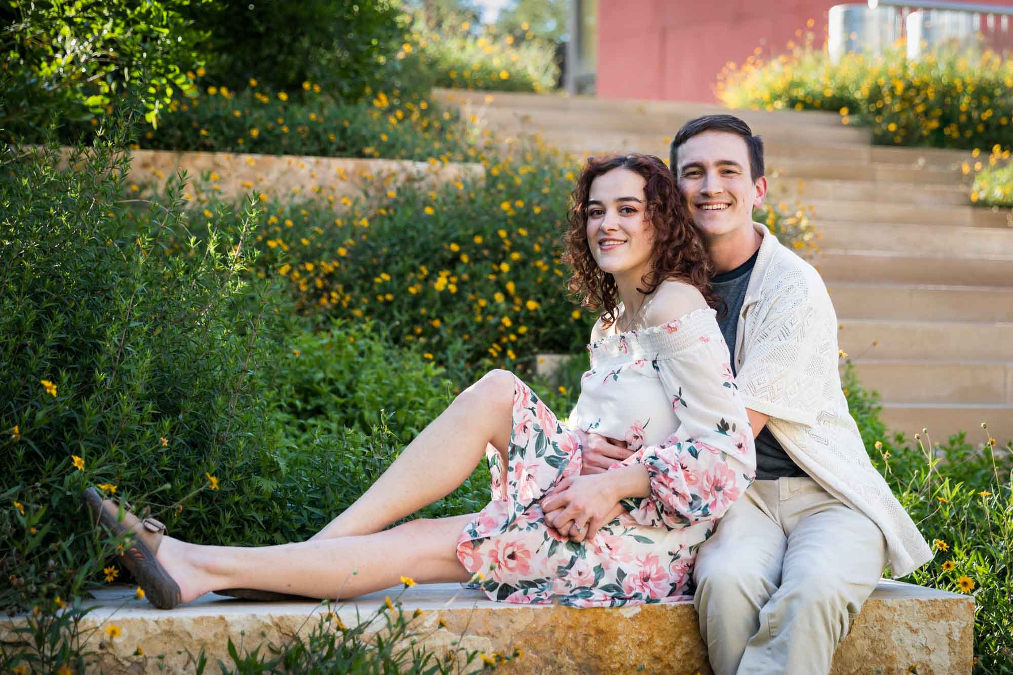 Downtown San Antonio engagement photos of couple seated on stone bench in front of green bushes in San Pedro Creek Park