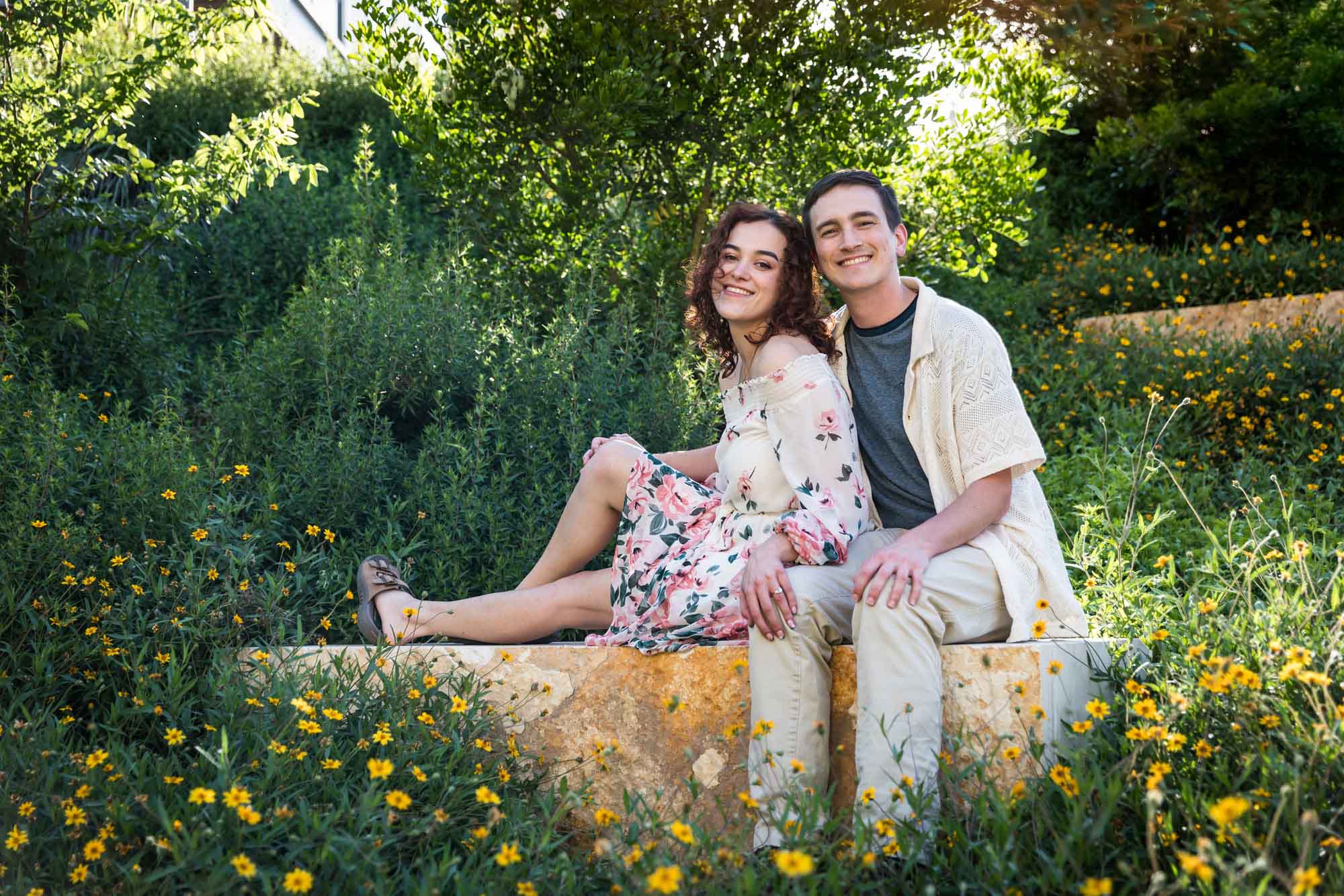 Downtown San Antonio engagement photos of couple seated on stone bench in front of green bushes in San Pedro Creek Park