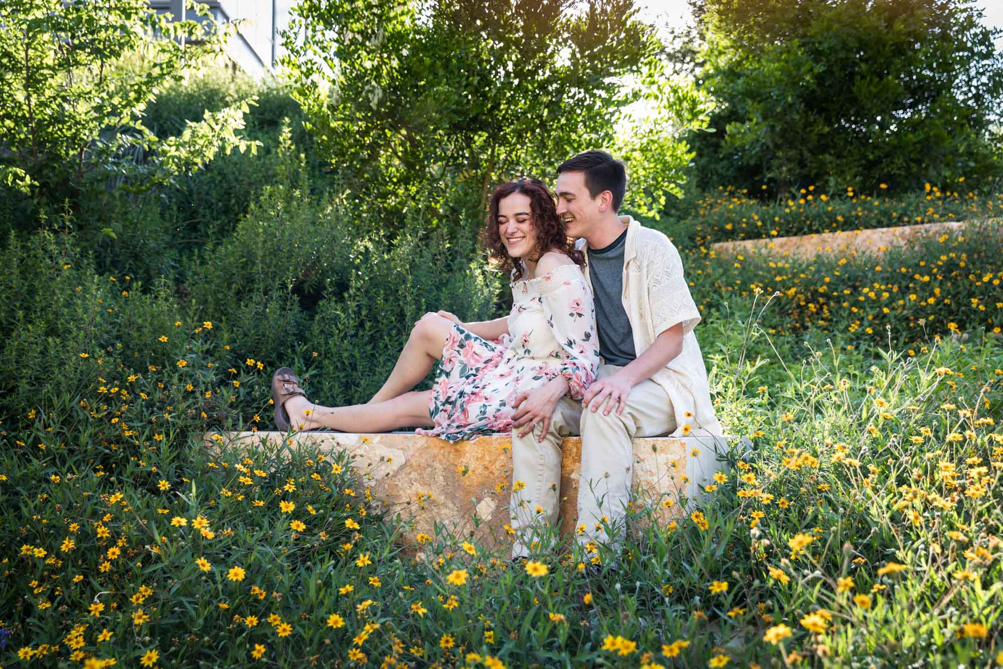 Downtown San Antonio engagement photos of couple seated on stone bench in front of green bushes in San Pedro Creek Park