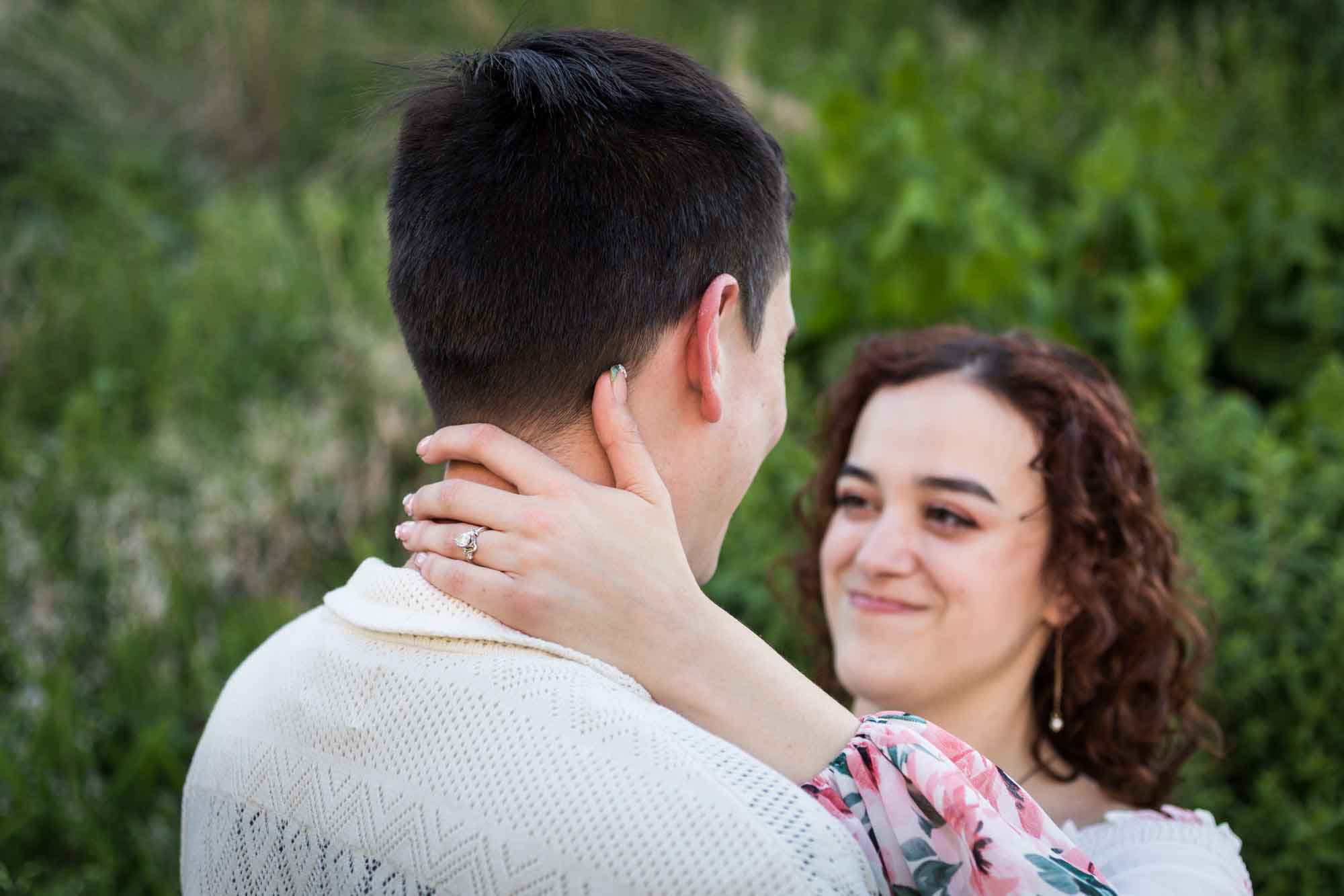 Downtown San Antonio engagement photos of woman with hand on man's neck showing engagement ring in front of green bushes in San Pedro Creek Park