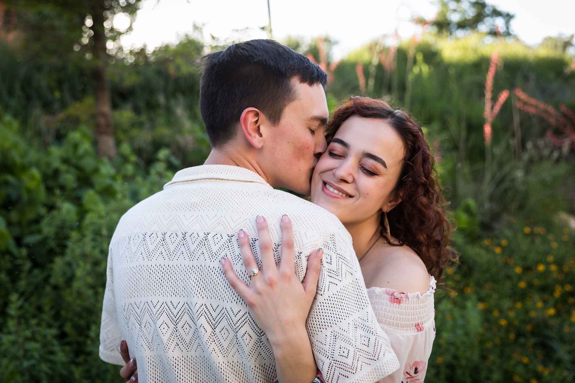 Downtown San Antonio engagement photos of man kissing woman on cheek in front of green bushes in San Pedro Creek Park