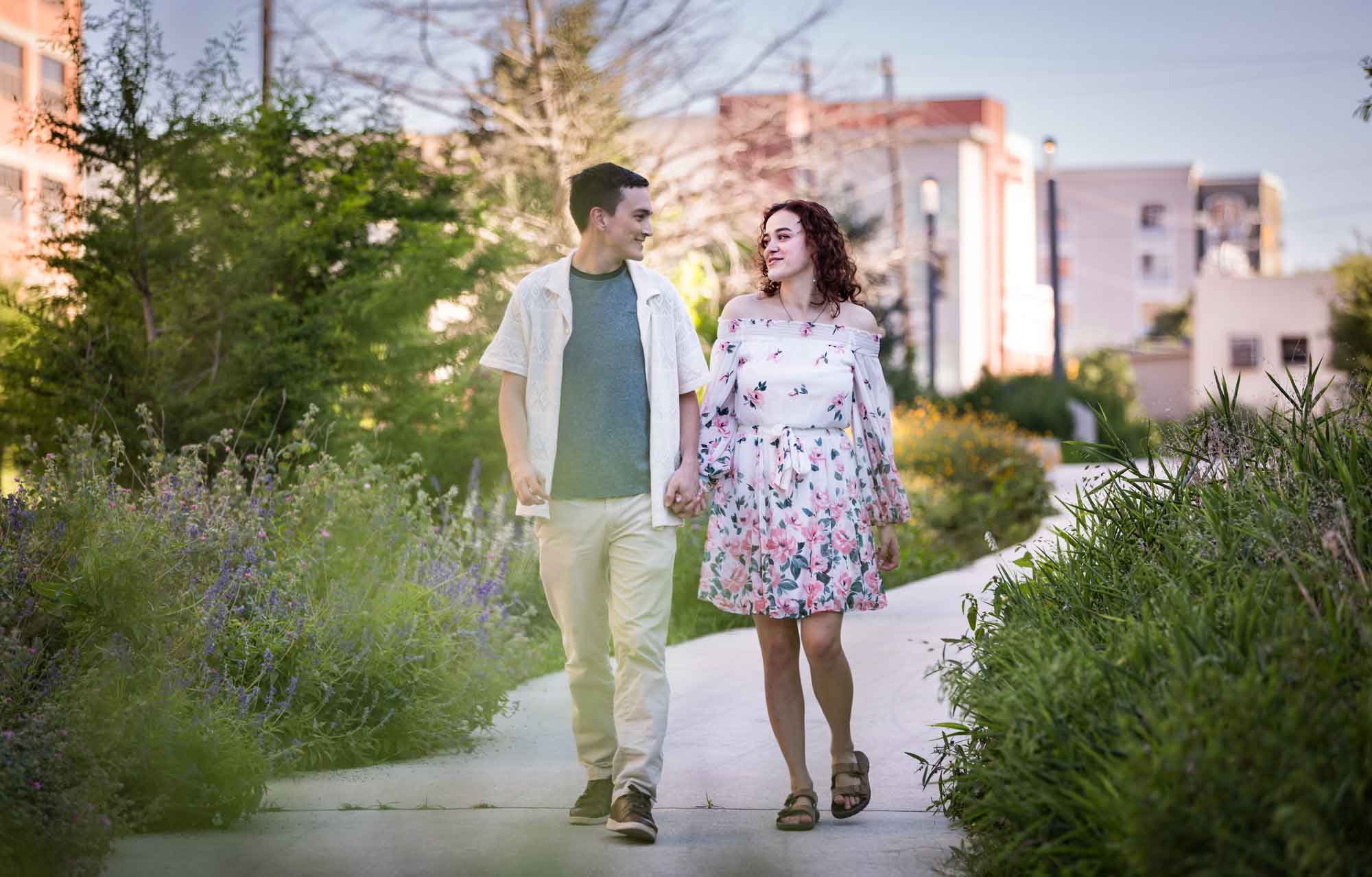 Downtown San Antonio engagement photos of couple walking in middle of pathway in front of green bushes in San Pedro Creek Park