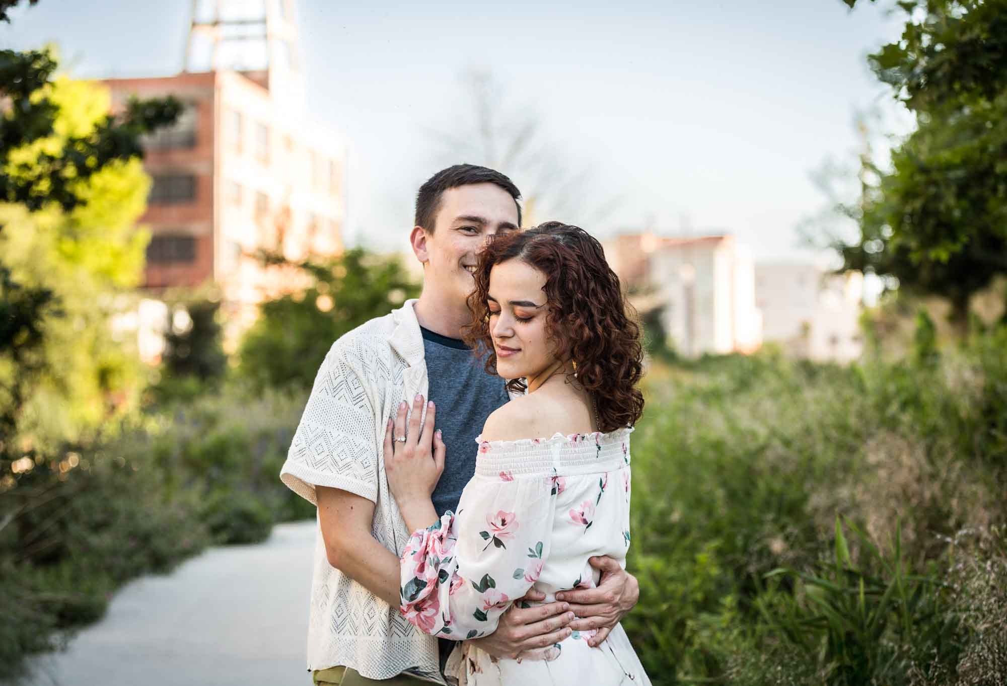 Downtown San Antonio engagement photos of couple hugging in middle of pathway in front of green bushes in San Pedro Creek Park