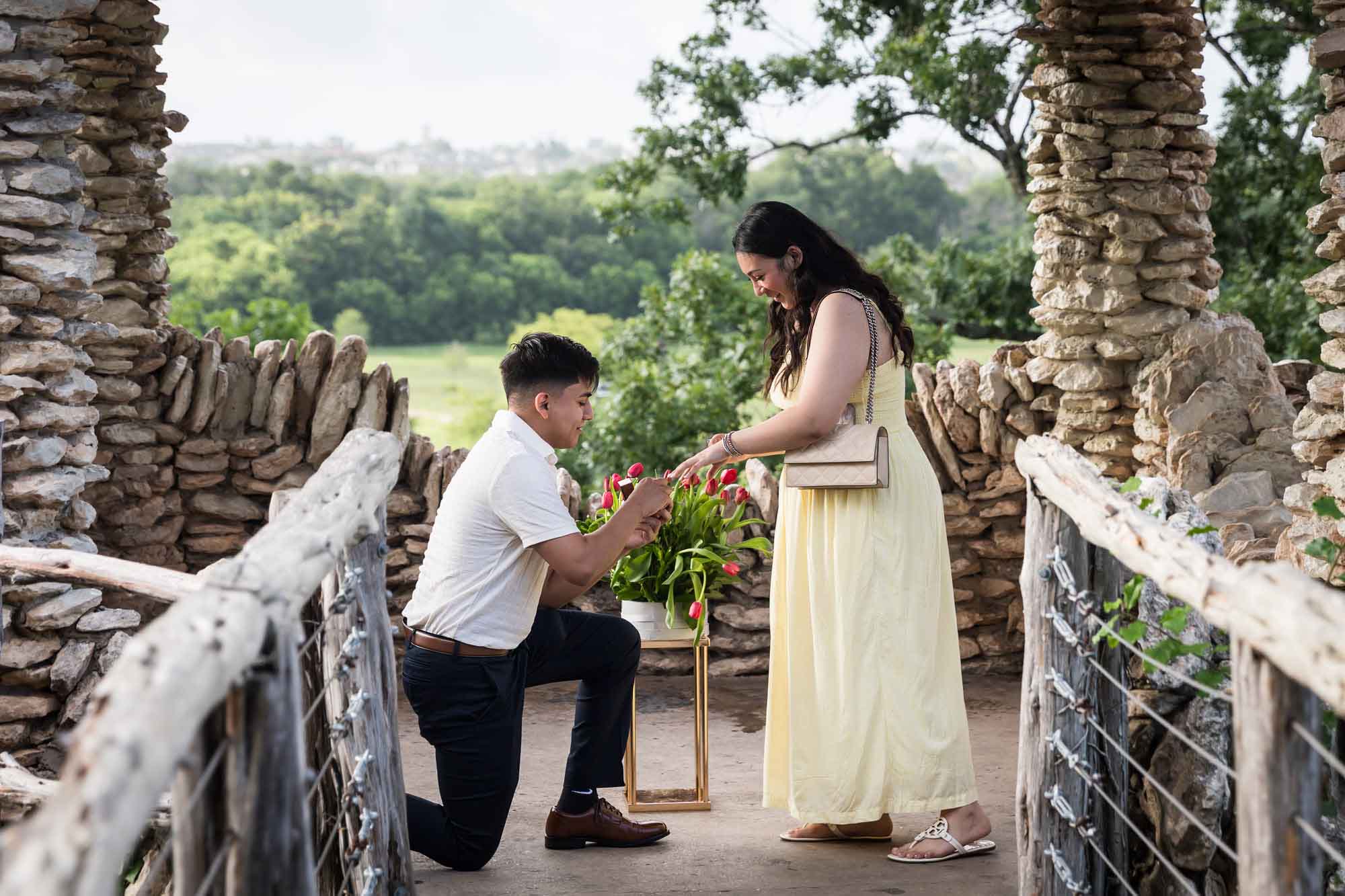Man wearing black pants bending down on one knee in front of woman wearing yellow dress in stone gazebo during proposal for an article on Japanese Tea Garden proposal tips