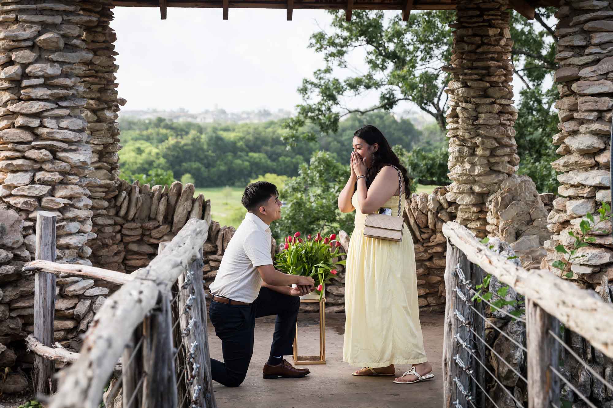 Man wearing black pants bending down on one knee in front of woman wearing yellow dress in stone gazebo during proposal for an article on Japanese Tea Garden proposal tips