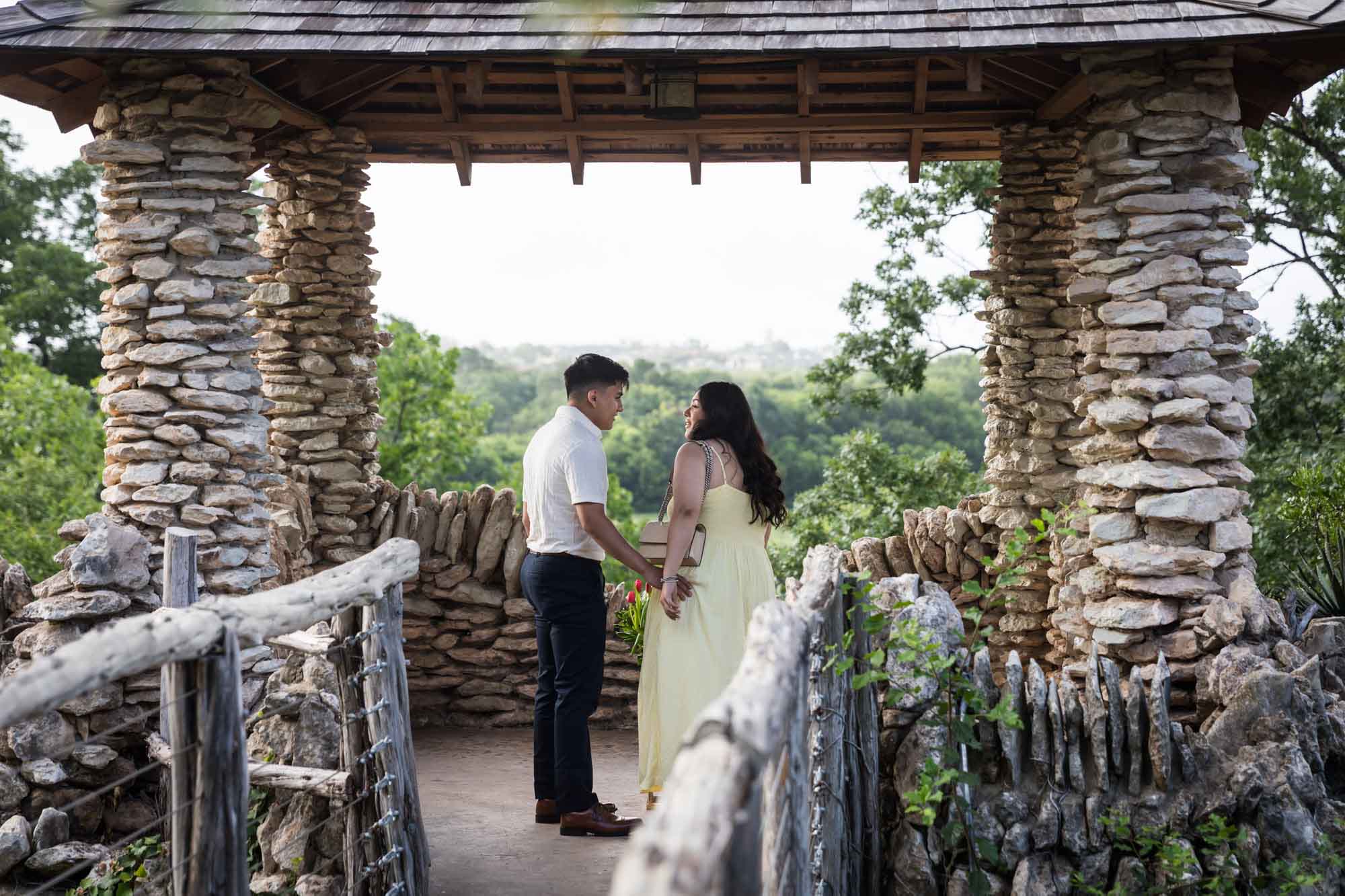 Man wearing black pants holding hands of woman wearing yellow dress in stone gazebo during proposal for an article on Japanese Tea Garden proposal tips
