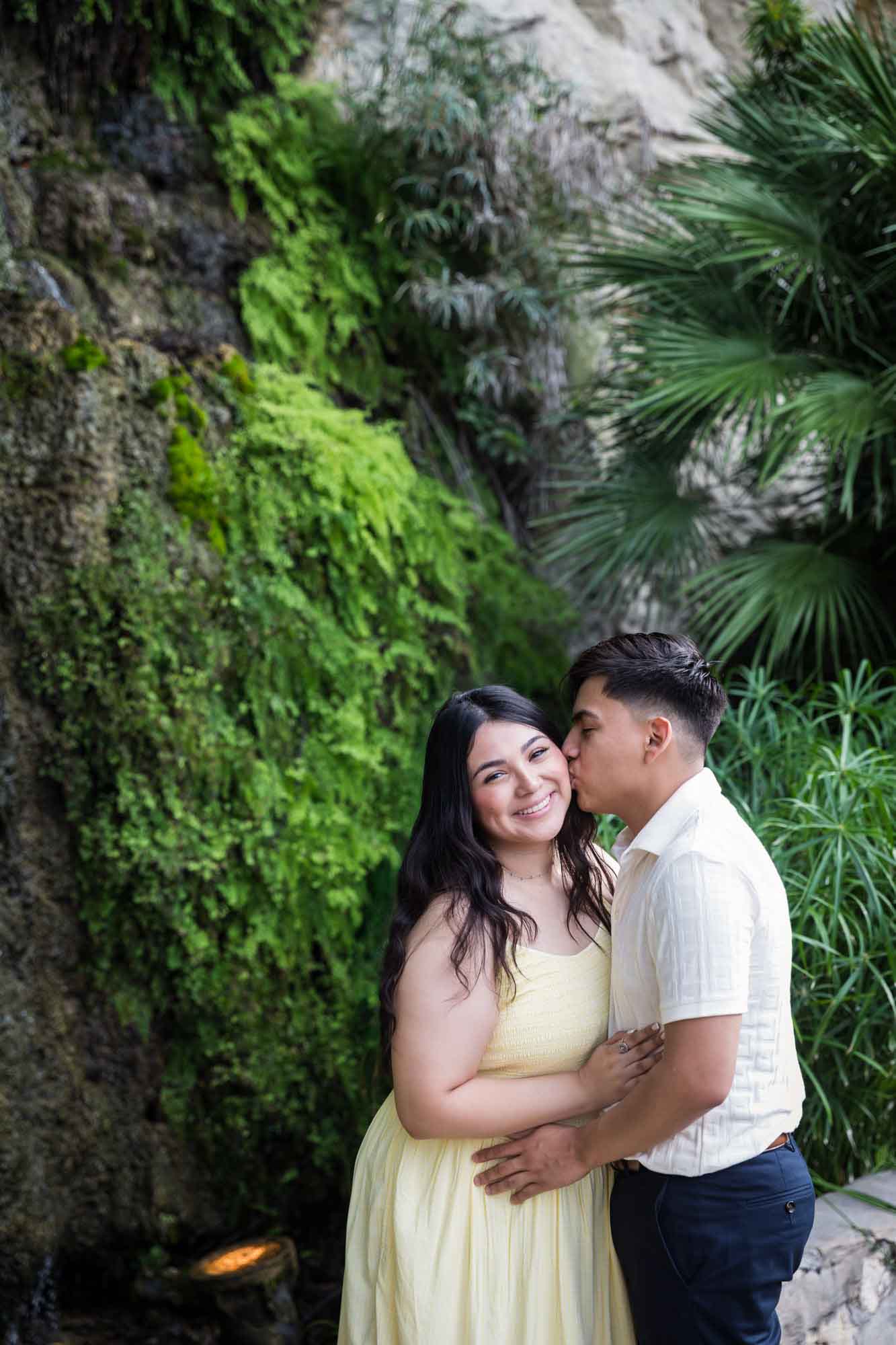Man wearing white shirt kissing woman wearing yellow dress in front of waterfall during a Japanese Tea Garden engagement portrait