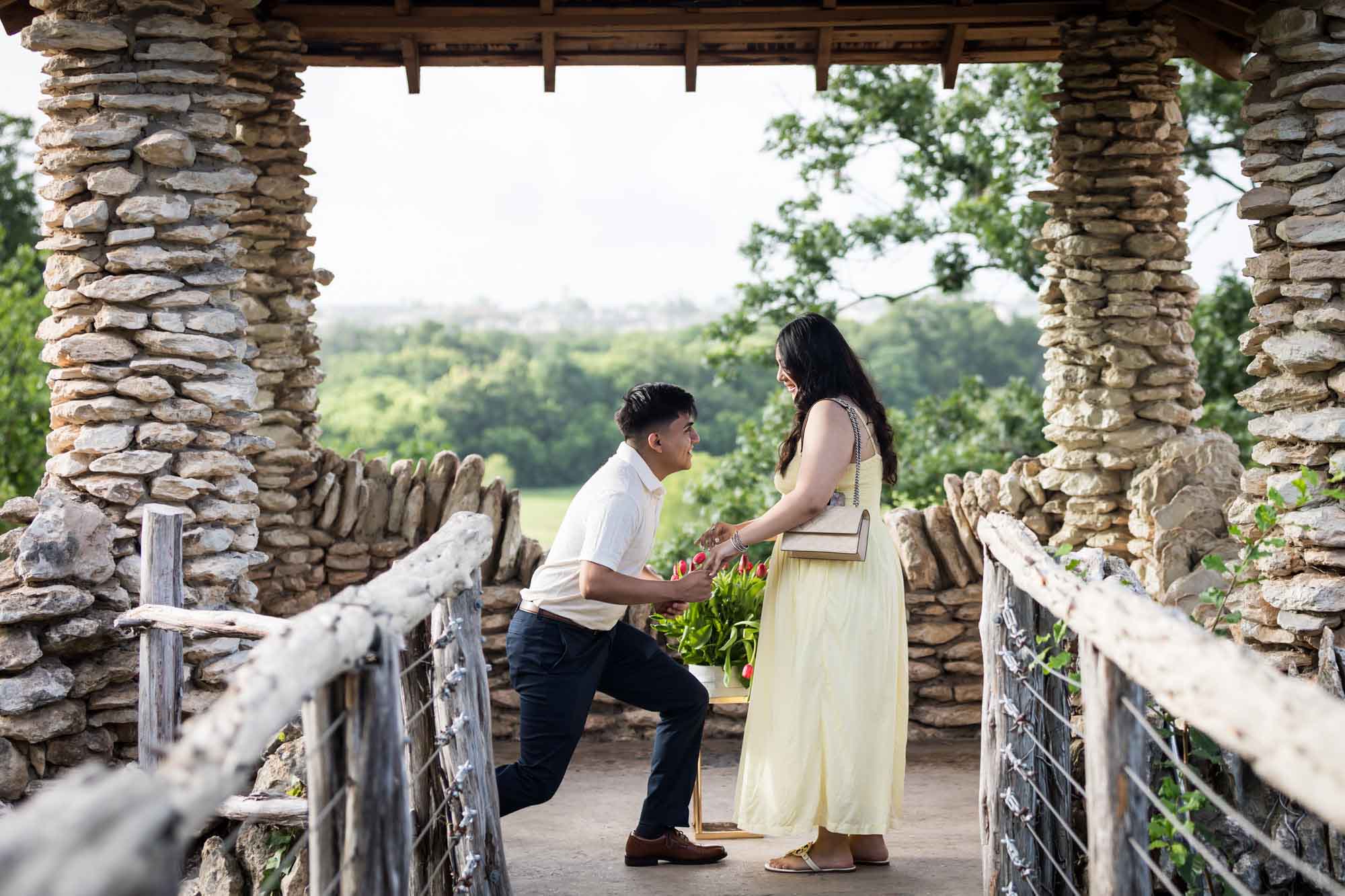 Man wearing black pants getting down on one knee in front of woman wearing yellow dress in stone gazebo during proposal for an article on Japanese Tea Garden proposal tips