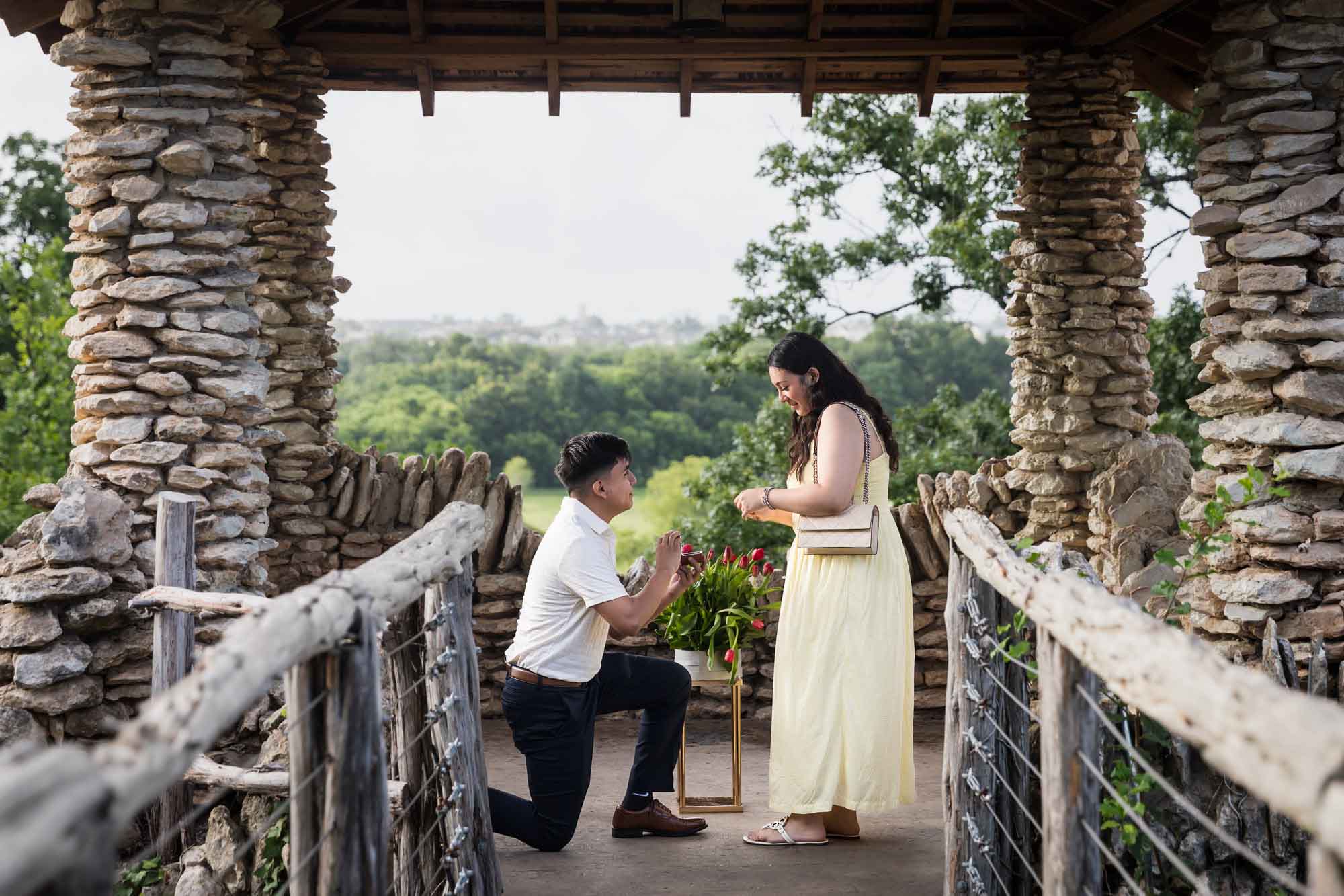 Man wearing black pants bending down on one knee in front of woman wearing yellow dress in stone gazebo during proposal for an article on Japanese Tea Garden proposal tips