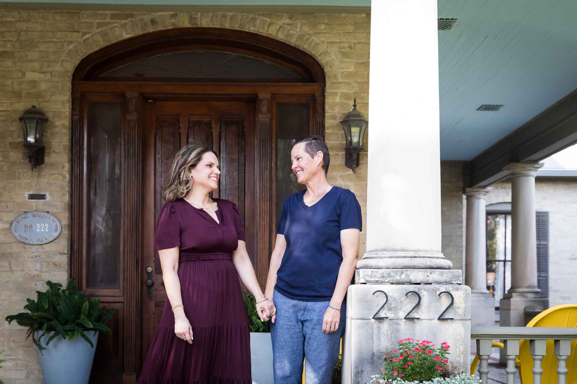 Two women standing side by side holding hands to the left of large pillar in front of house during a King William family portrait