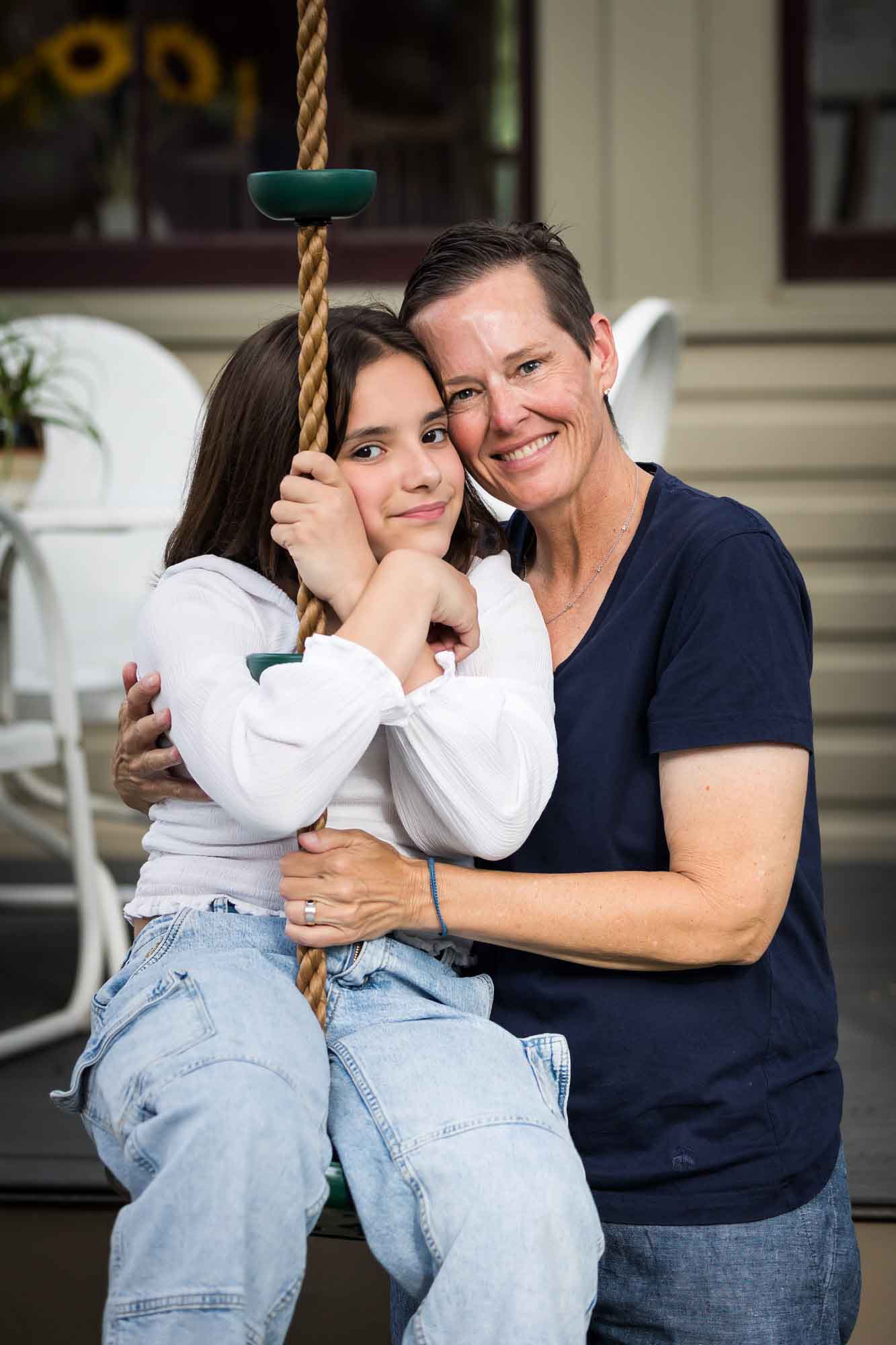 Mother wearing navy t-shirt hugging daughter wearing white shirt playing on swing Mother wearing maroon dress seated with son wearing blue shirt and dog Teenage girl wearing white blouse and jeans standing on swing during a King William family portrait