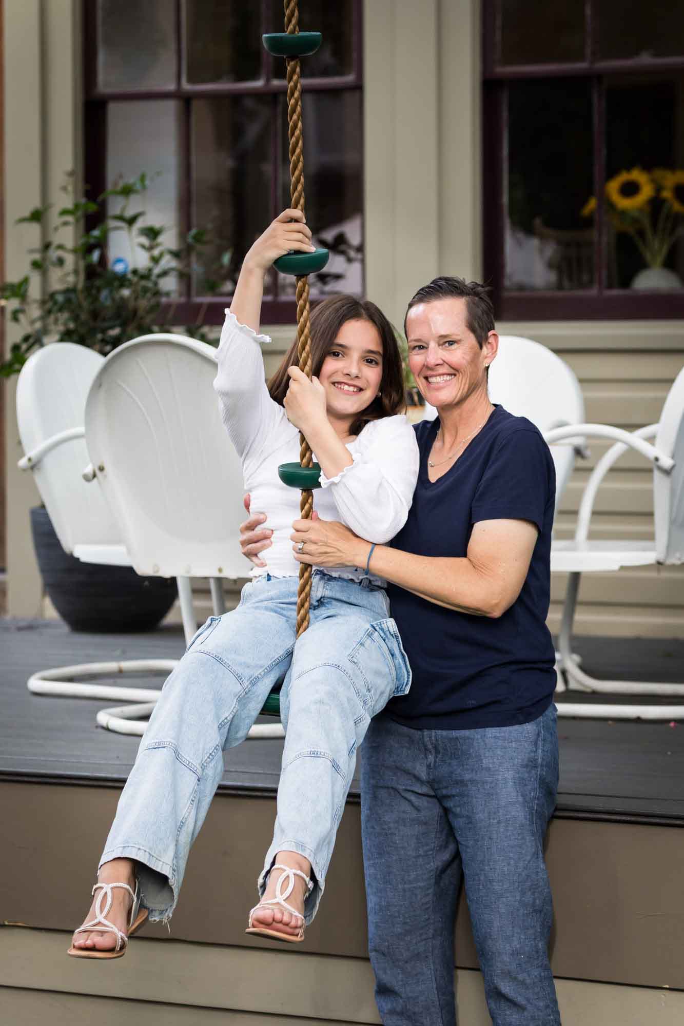 Mother wearing navy t-shirt hugging daughter wearing white shirt playing on swing Mother wearing maroon dress seated with son wearing blue shirt and dog Teenage girl wearing white blouse and jeans standing on swing during a King William family portrait