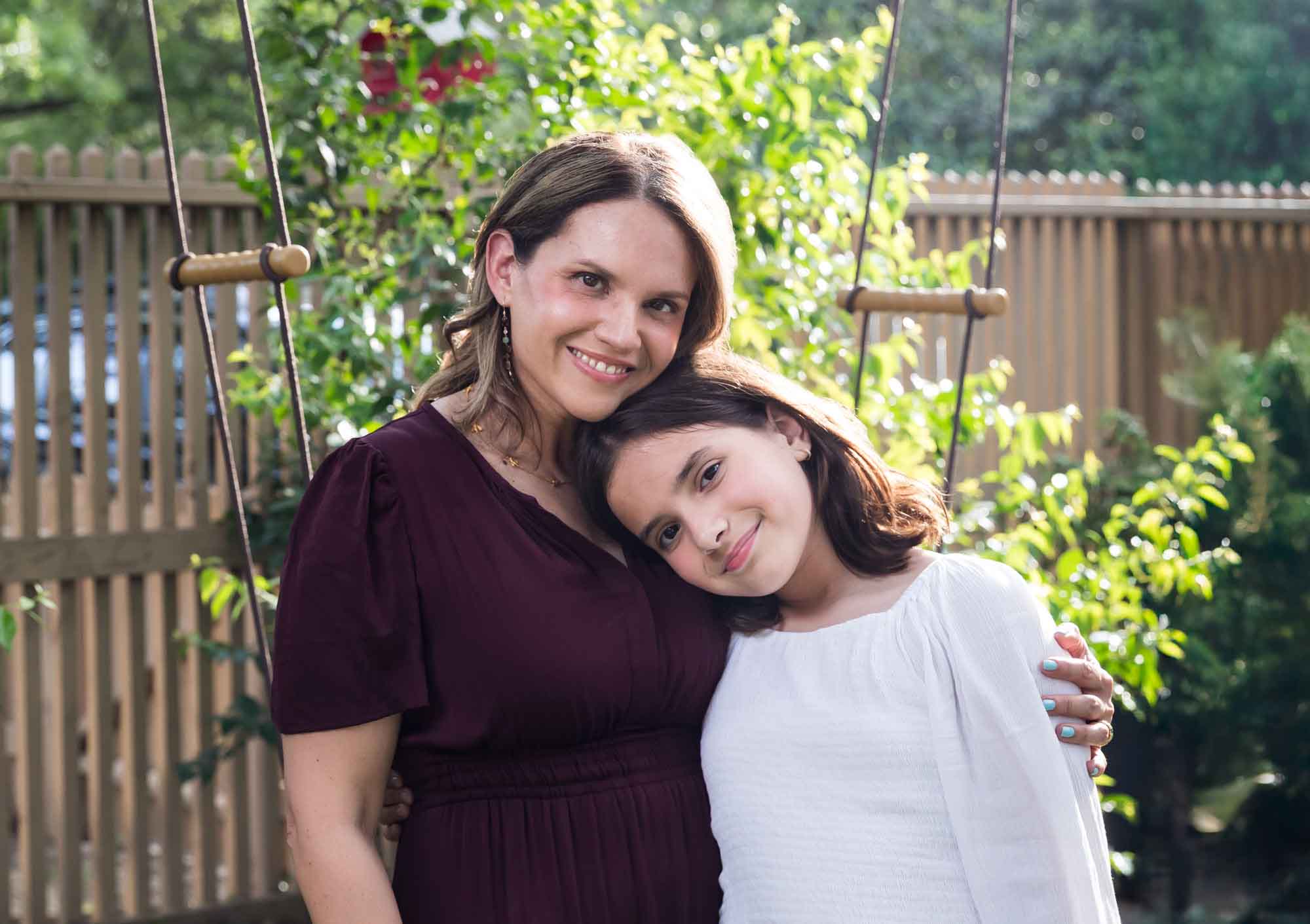 Mother wearing maroon dress hugging teenage daughter wearing white shirt in backyard Mother wearing navy t-shirt hugging daughter wearing white shirt playing on swing Mother wearing maroon dress seated with son wearing blue shirt and dog Teenage girl wearing white blouse and jeans standing on swing during a King William family portrait