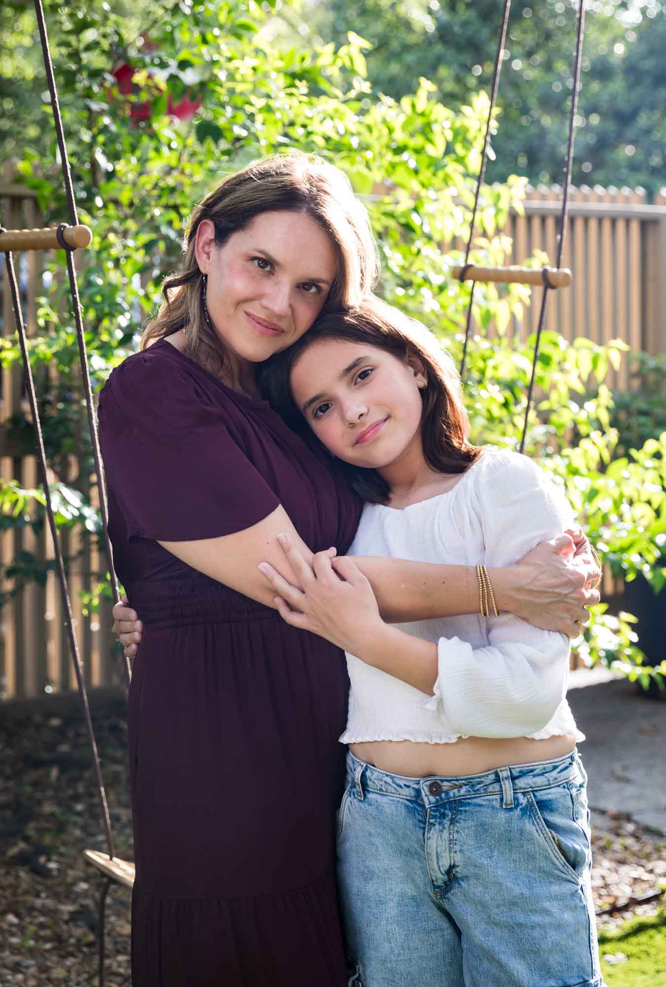 Mother wearing maroon dress hugging teenage daughter wearing white shirt and jeans in backyard Mother wearing navy t-shirt hugging daughter wearing white shirt playing on swing Mother wearing maroon dress seated with son wearing blue shirt and dog Teenage girl wearing white blouse and jeans standing on swing during a King William family portrait