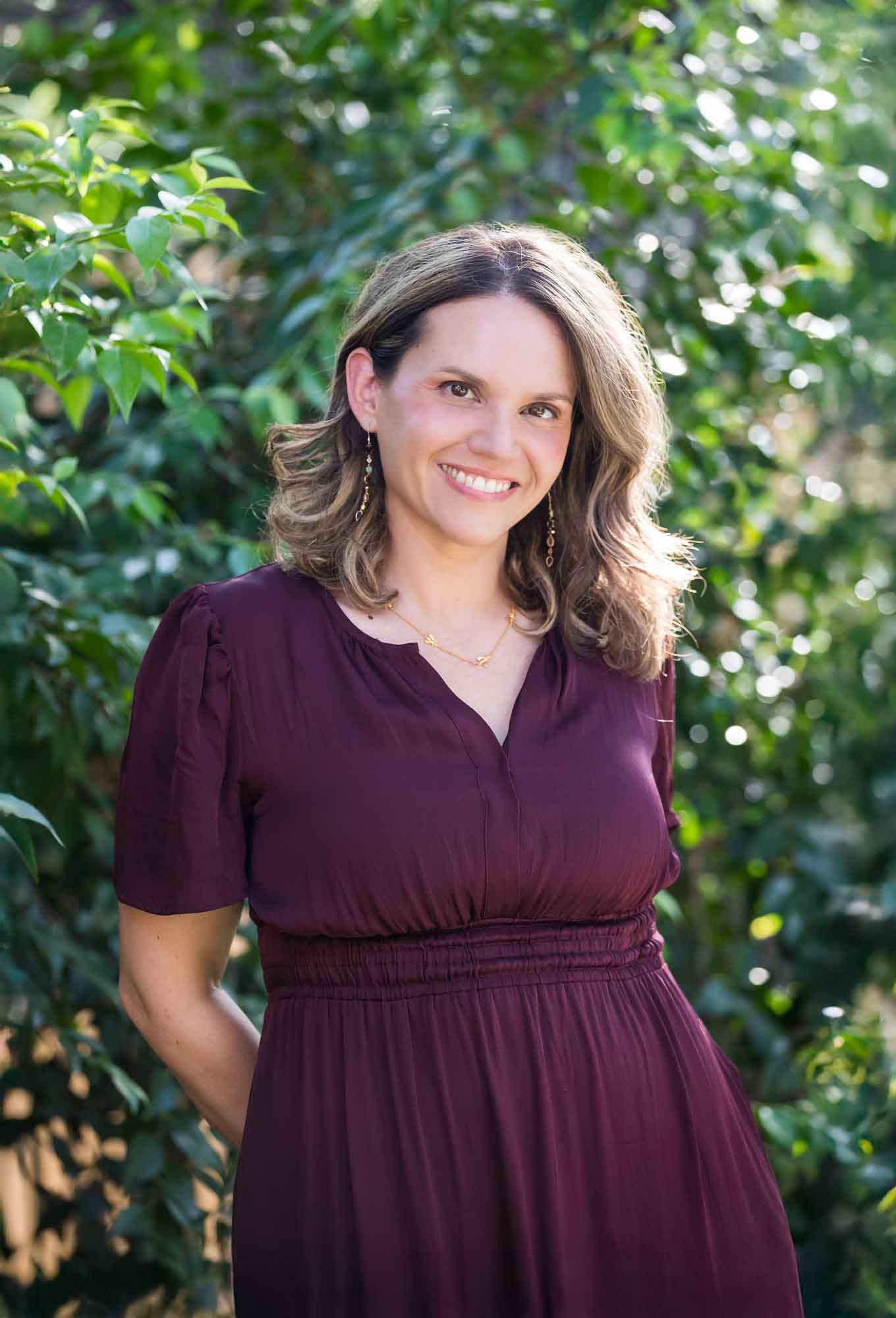 Woman wearing maroon dress with medium length brown hair standing in front of green bushes
