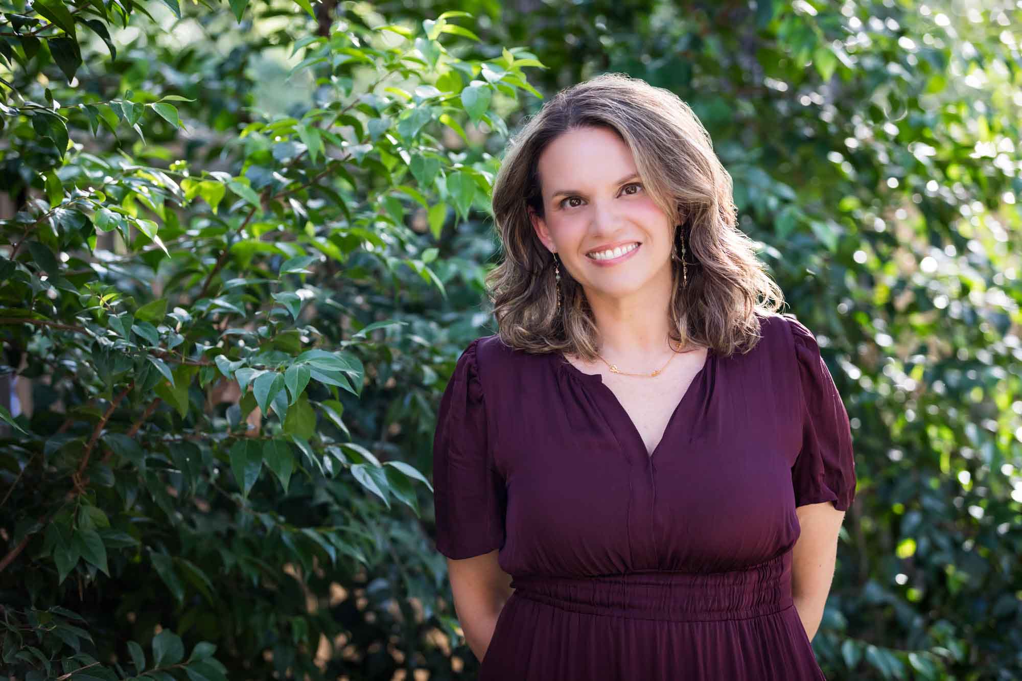 Woman wearing maroon dress with medium length brown hair standing in front of green bushes