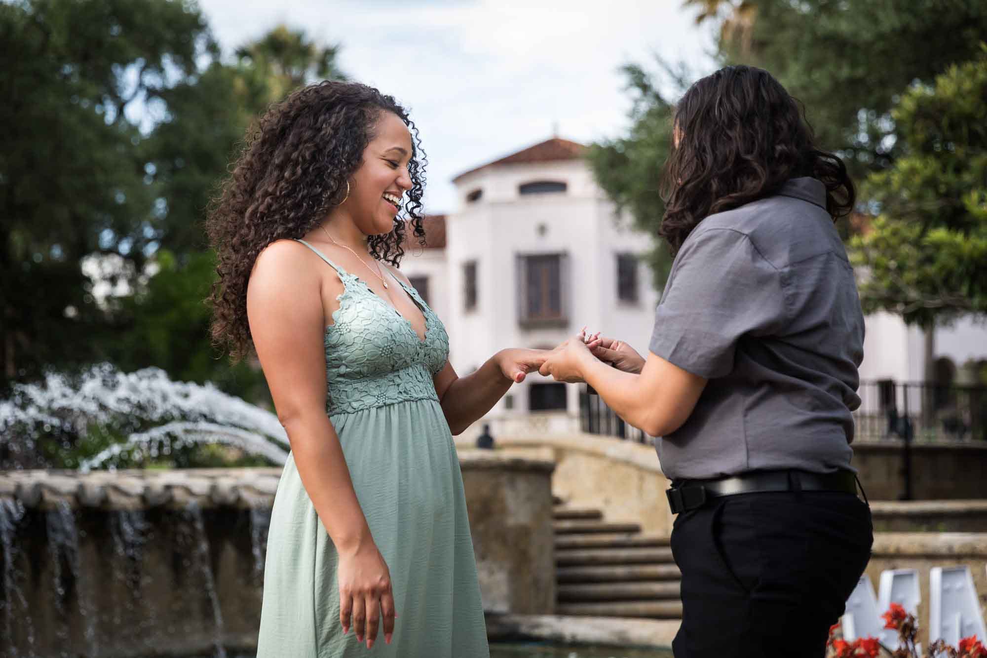 Woman putting engagement ring on another woman's hand in front of fountain for an article about McNay Art Museum proposal tips