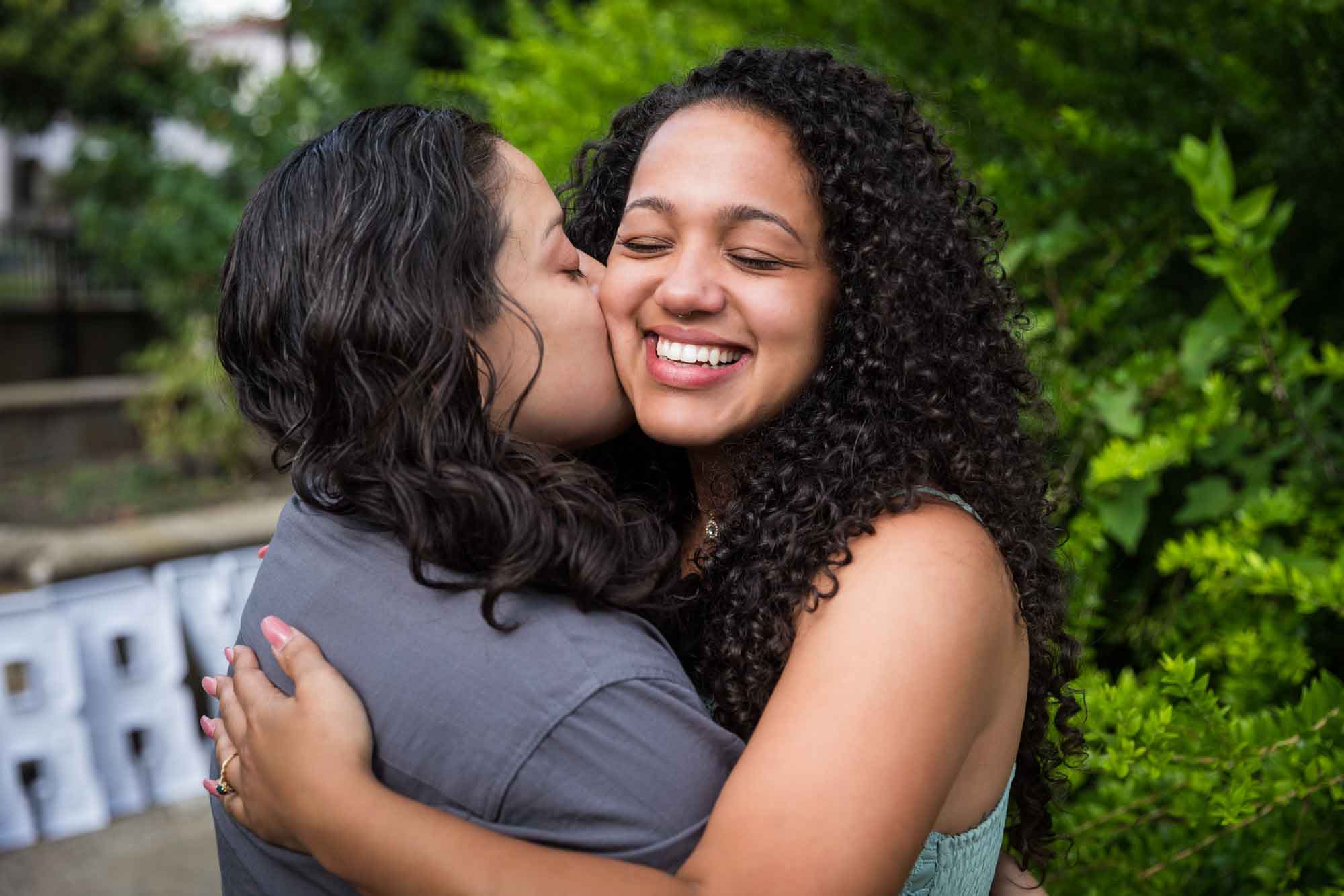 Woman kissing another woman on the cheek in front of green bush