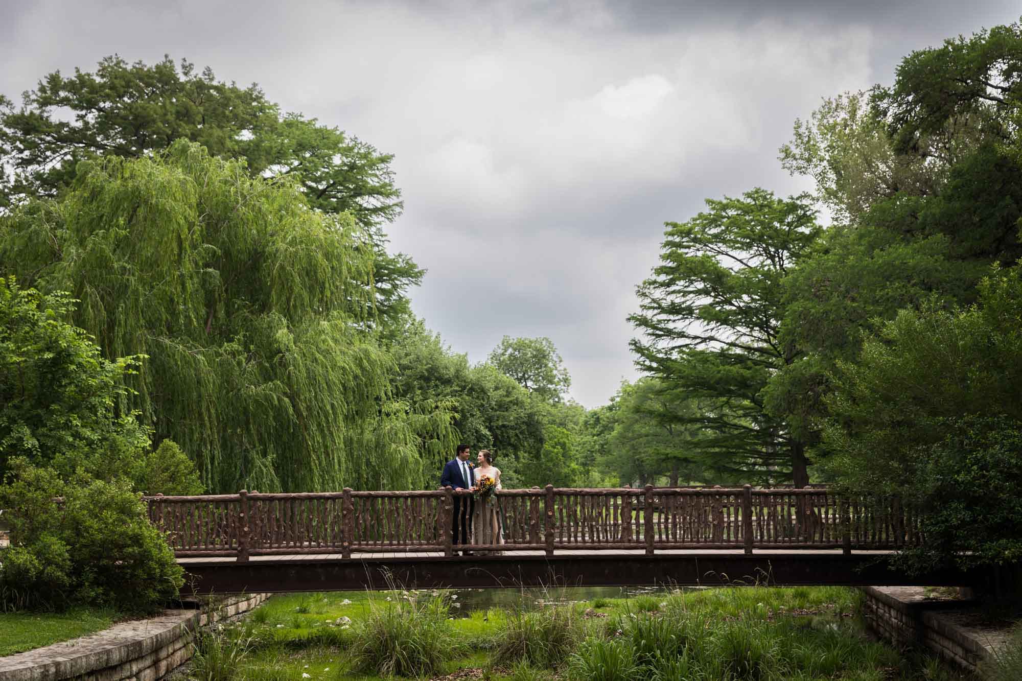 Bride and groom standing together on wooden bridge surrounded by trees in Landa Park for an article on affordable wedding venues in San Antonio
