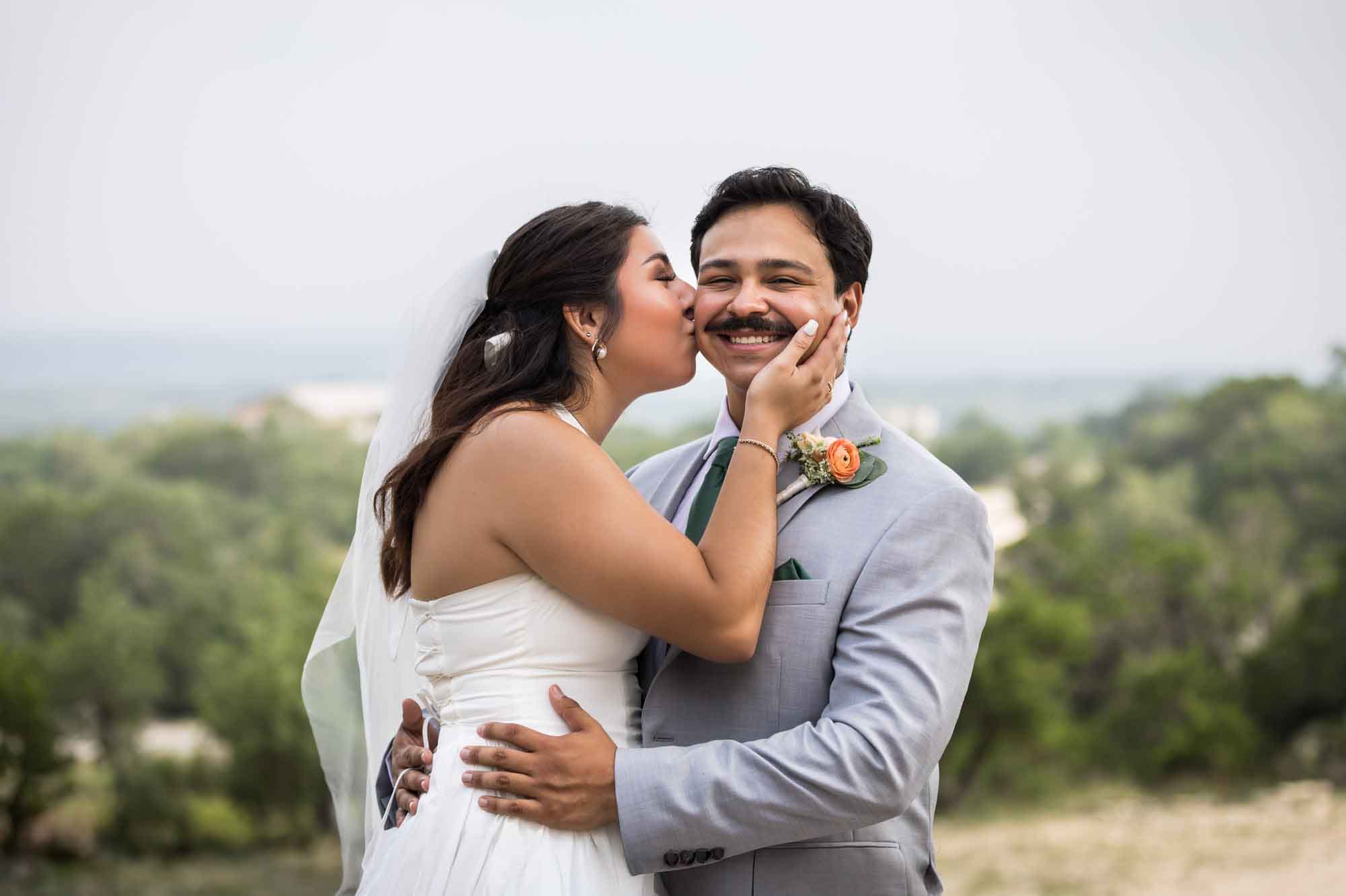 Bride kissing groom on the cheek with hill country view in the background for an article on affordable wedding venues in San Antonio