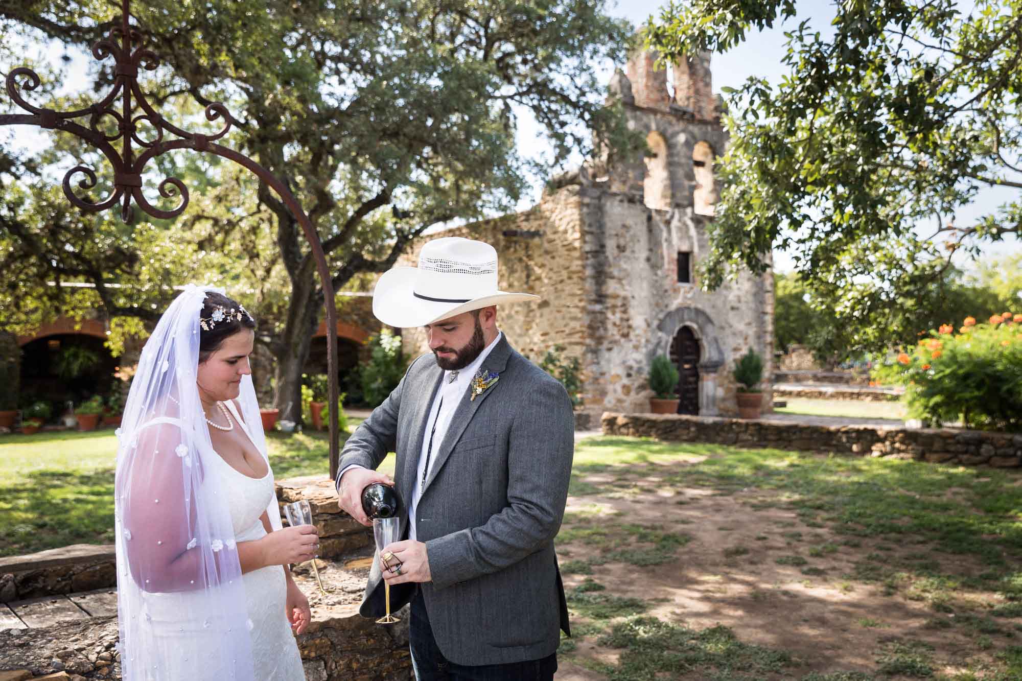 Bride and groom wearing cowboy hat pouring champagne with Mission Espada in background for an article on affordable wedding venues in San Antonio