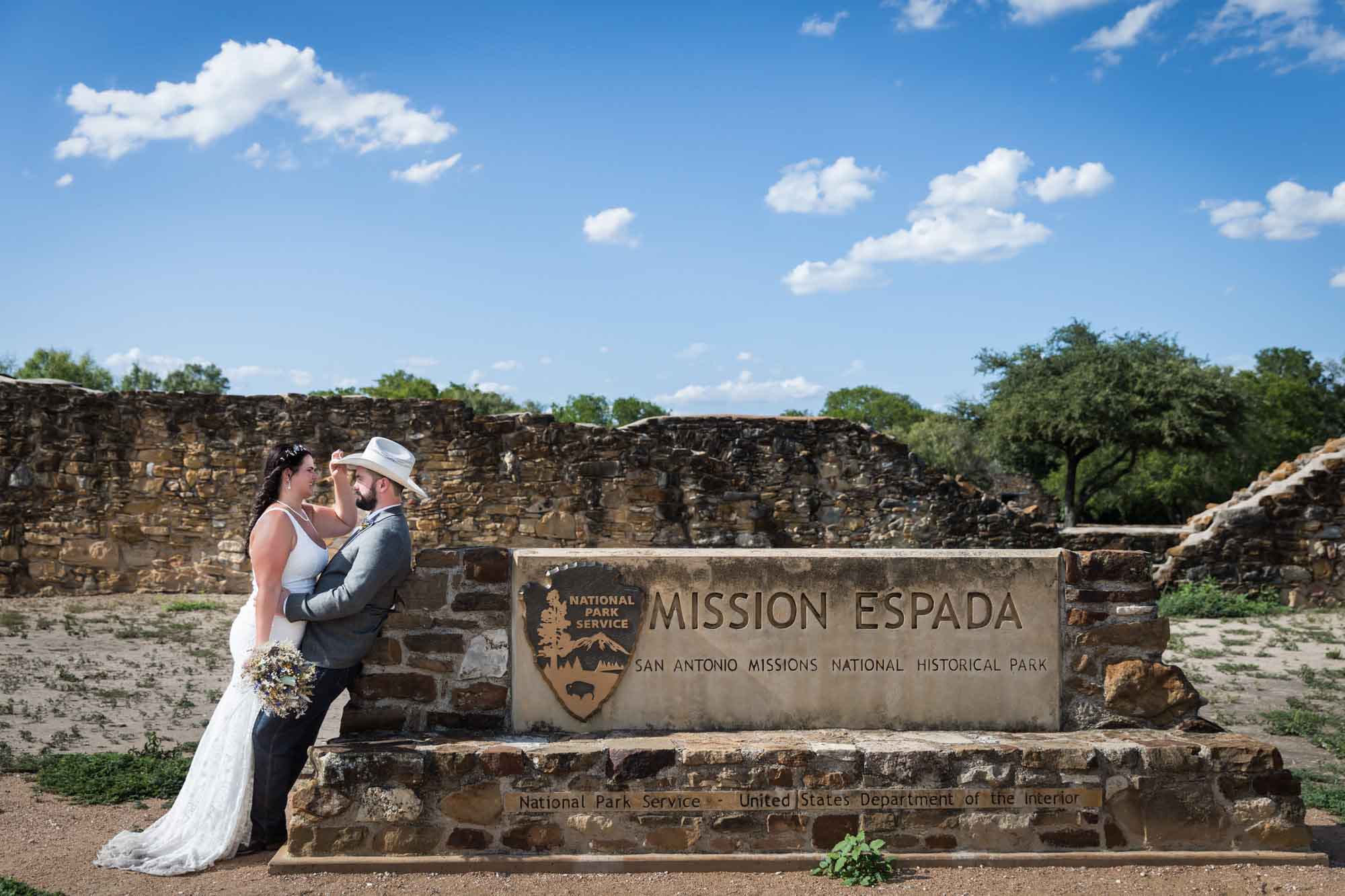 Bride and groom wearing cowboy hat leaning on stone sign for Mission Espada with blue sky overhead for an article on affordable wedding venues in San Antonio