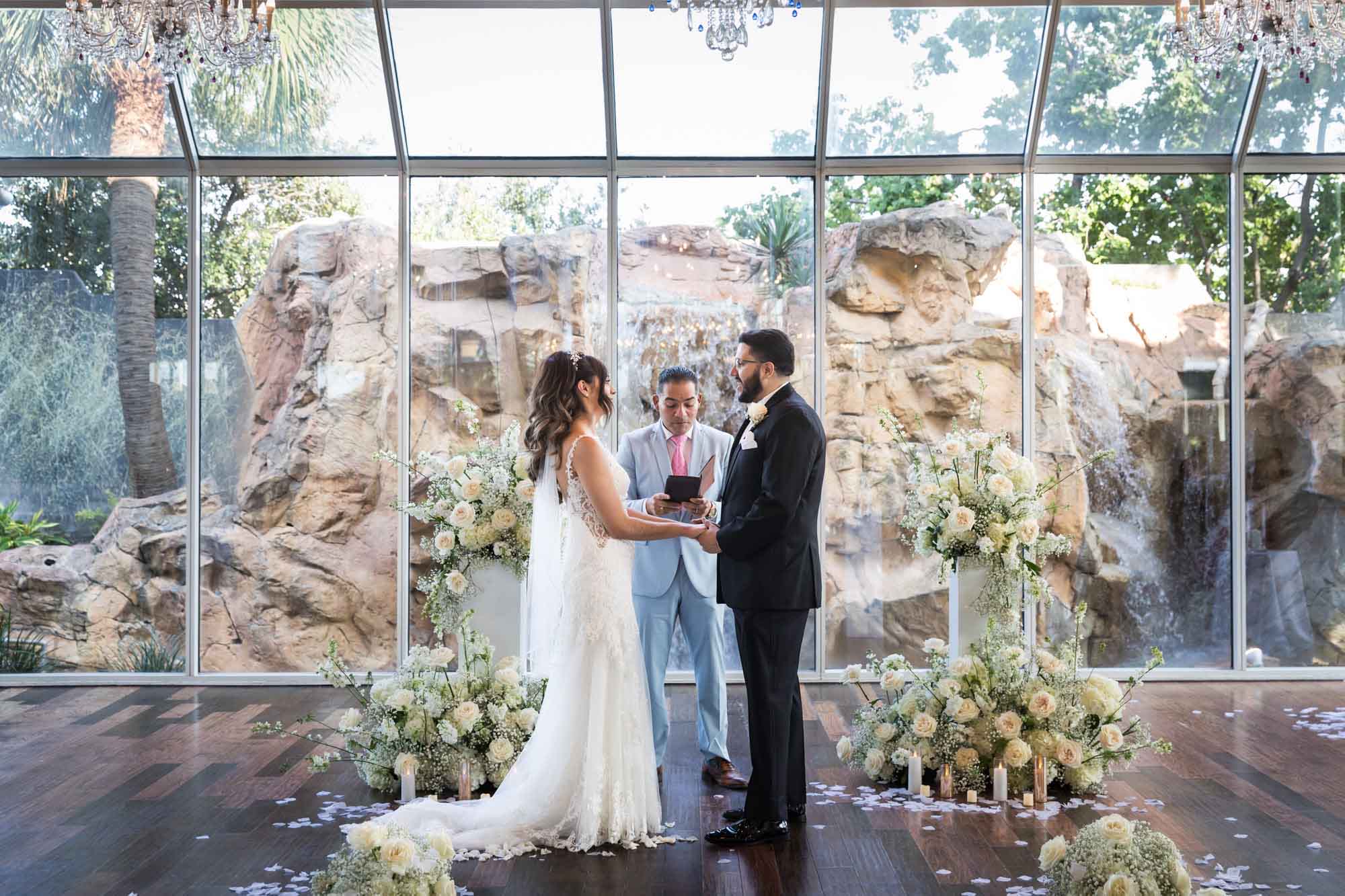 Bride and groom saying vows with officiant in front of glass wall and bouquets of white flowers for an article on affordable wedding venues in San Antonio