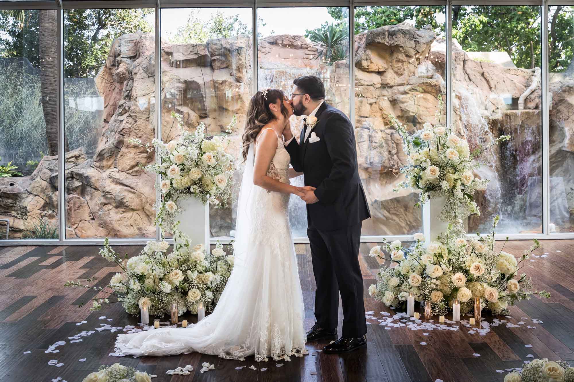 Bride and groom kissing in front of glass wall and bouquets of white flowers for an article on affordable wedding venues in San Antonio