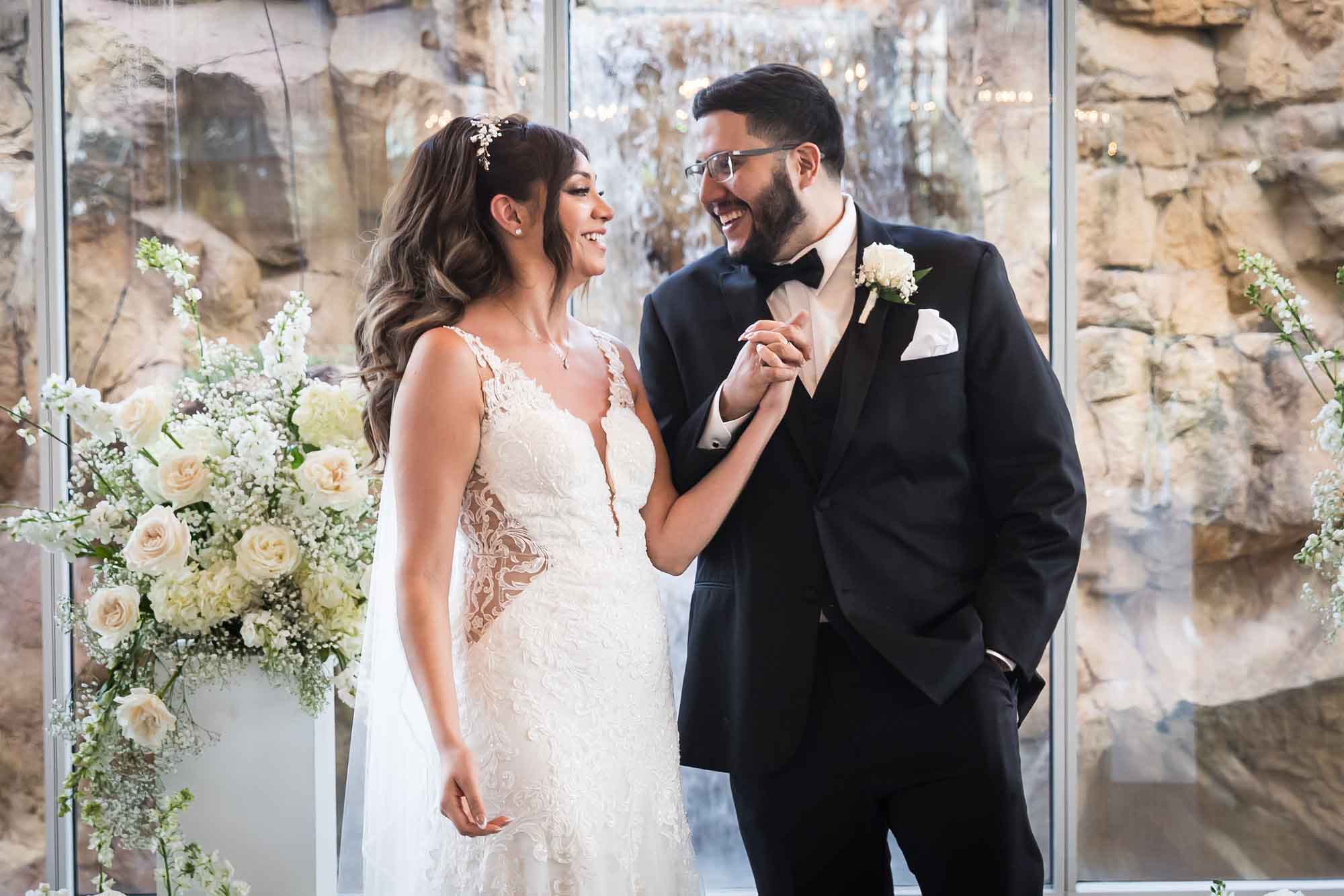 Groom holding bride's hand while standing in front of glass wall and white flowers