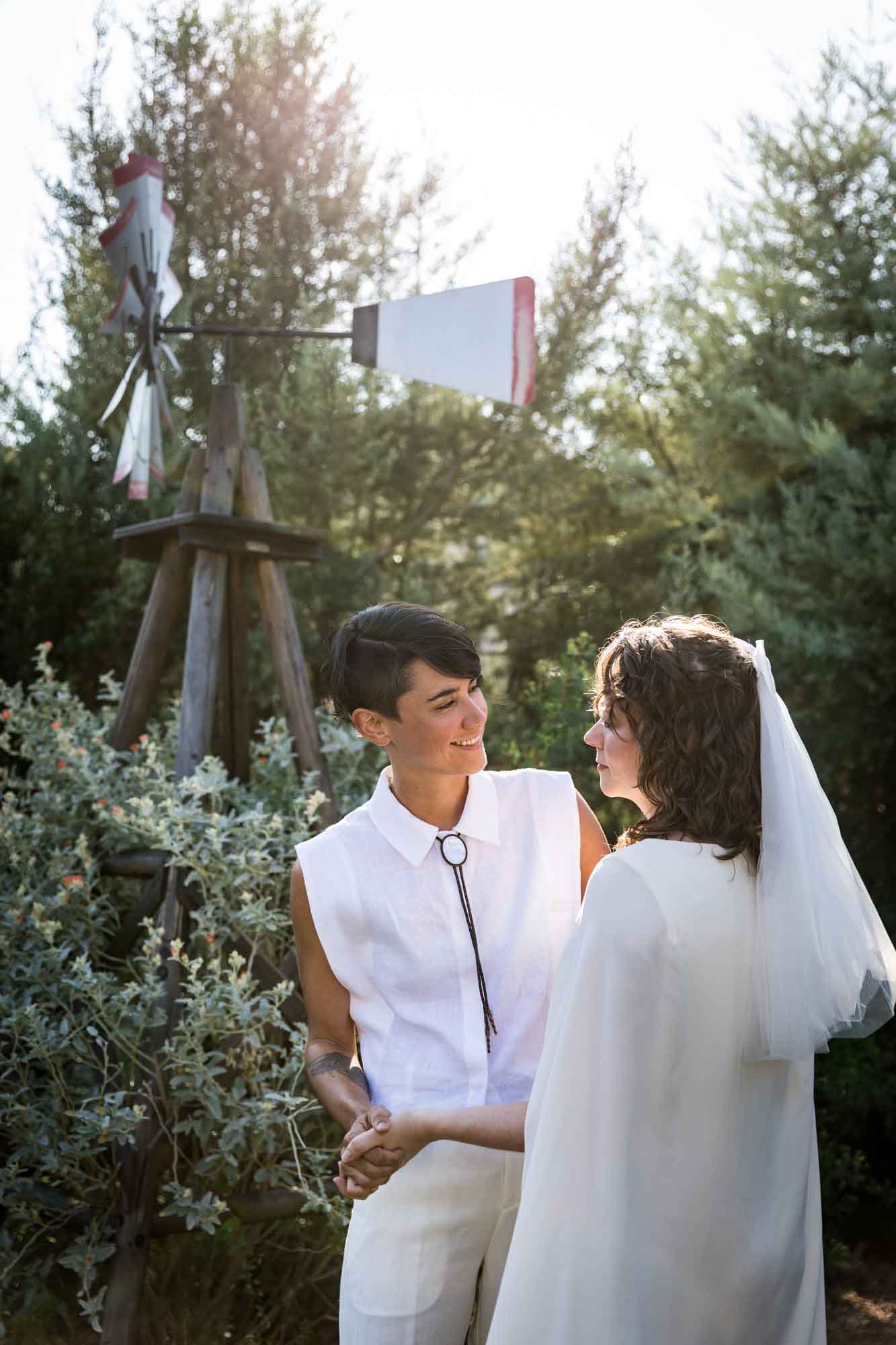 Two brides smiling at each other in front of large windmill in backyard for an article on affordable wedding venues in San Antonio