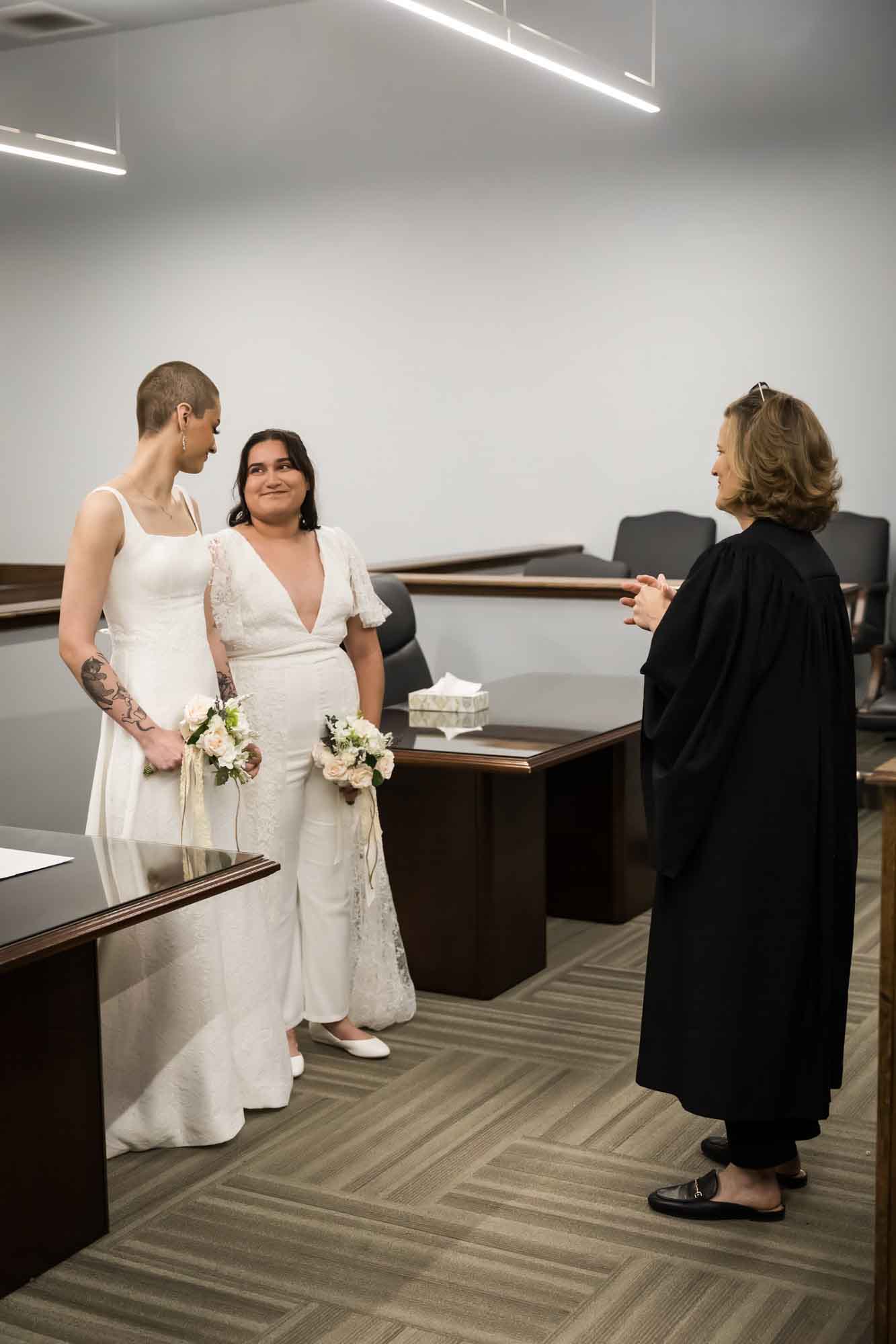 Two brides wearing white dresses looking at each other standing in front of a female judge for an article on affordable wedding venues in San Antonio