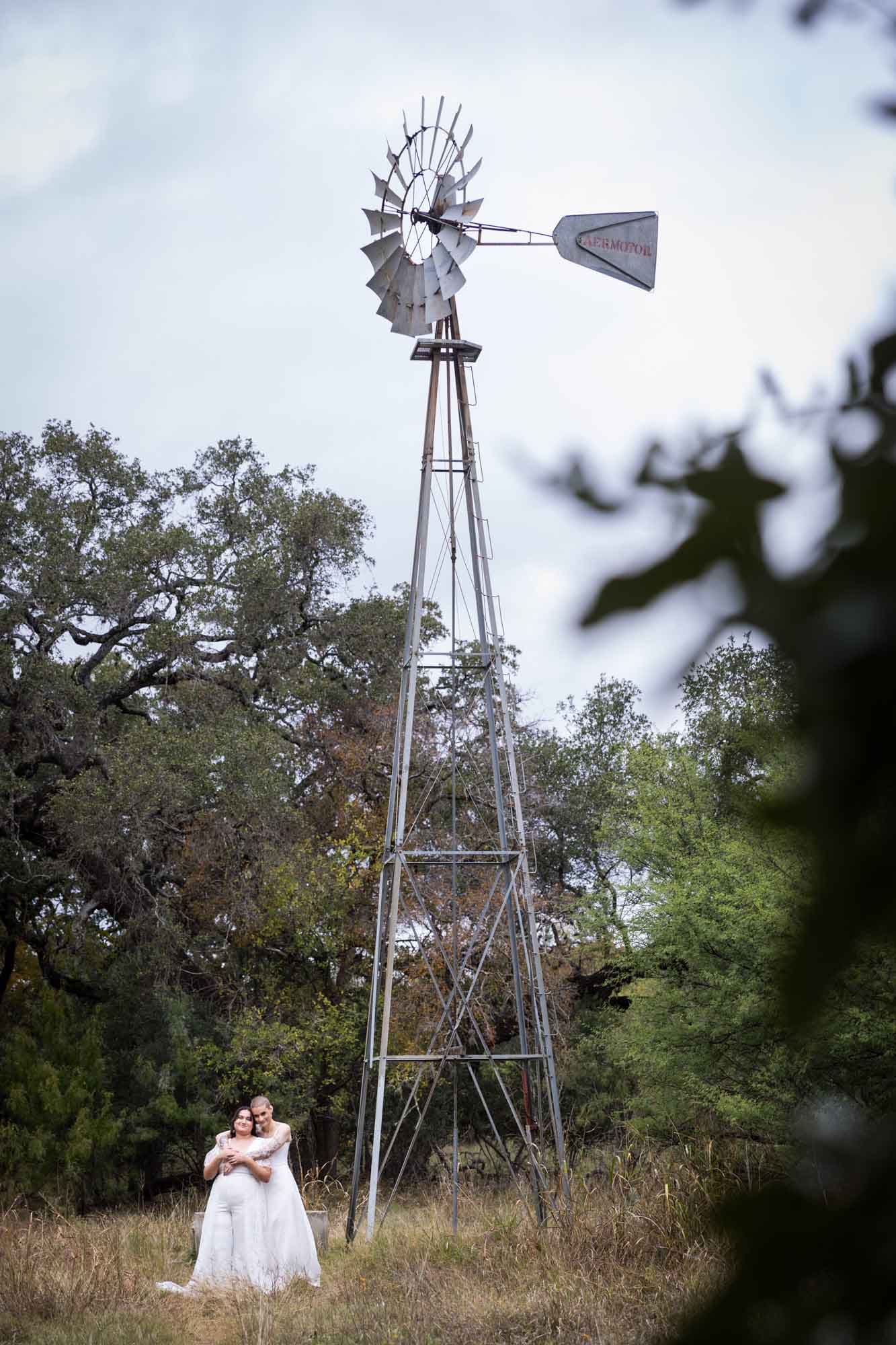 Two brides hugging at the base of a large windmill at Walker Ranch Park for an article on affordable wedding venues in San Antonio