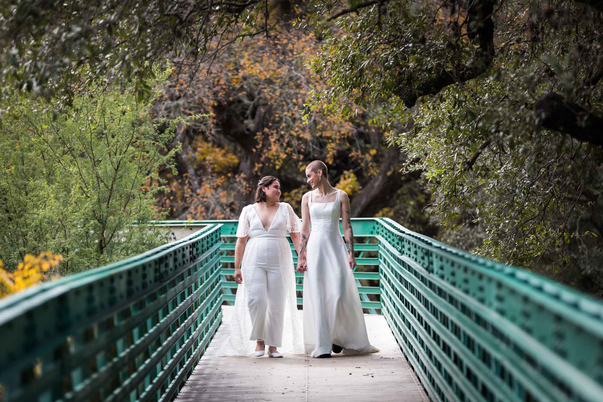 Two brides holding hands and walking over a green bridge at Walker Ranch Park for an article on affordable wedding venues in San Antonio