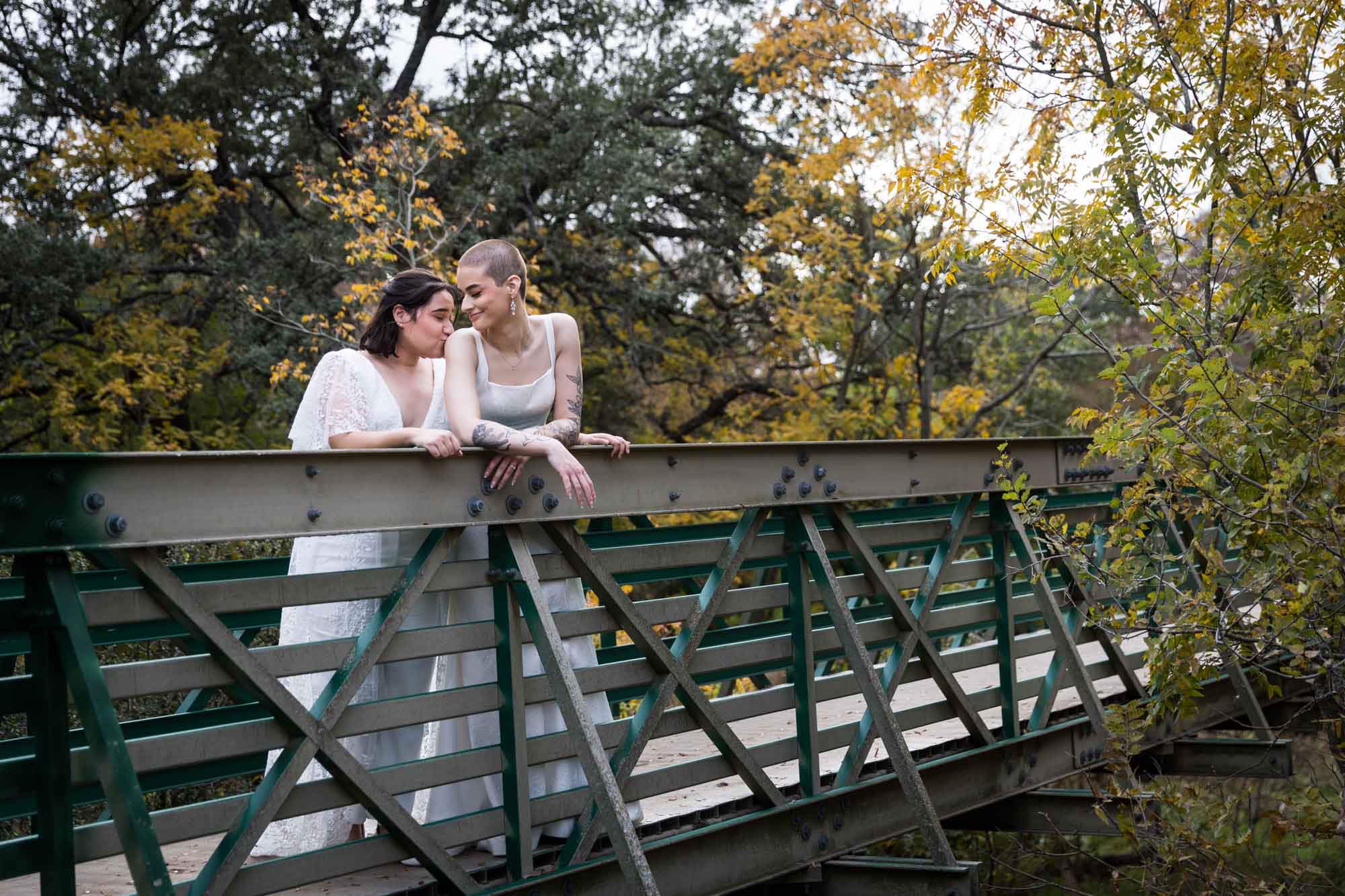 Two brides leaning on a green bridge railing at Walker Ranch Park for an article on affordable wedding venues in San Antonio