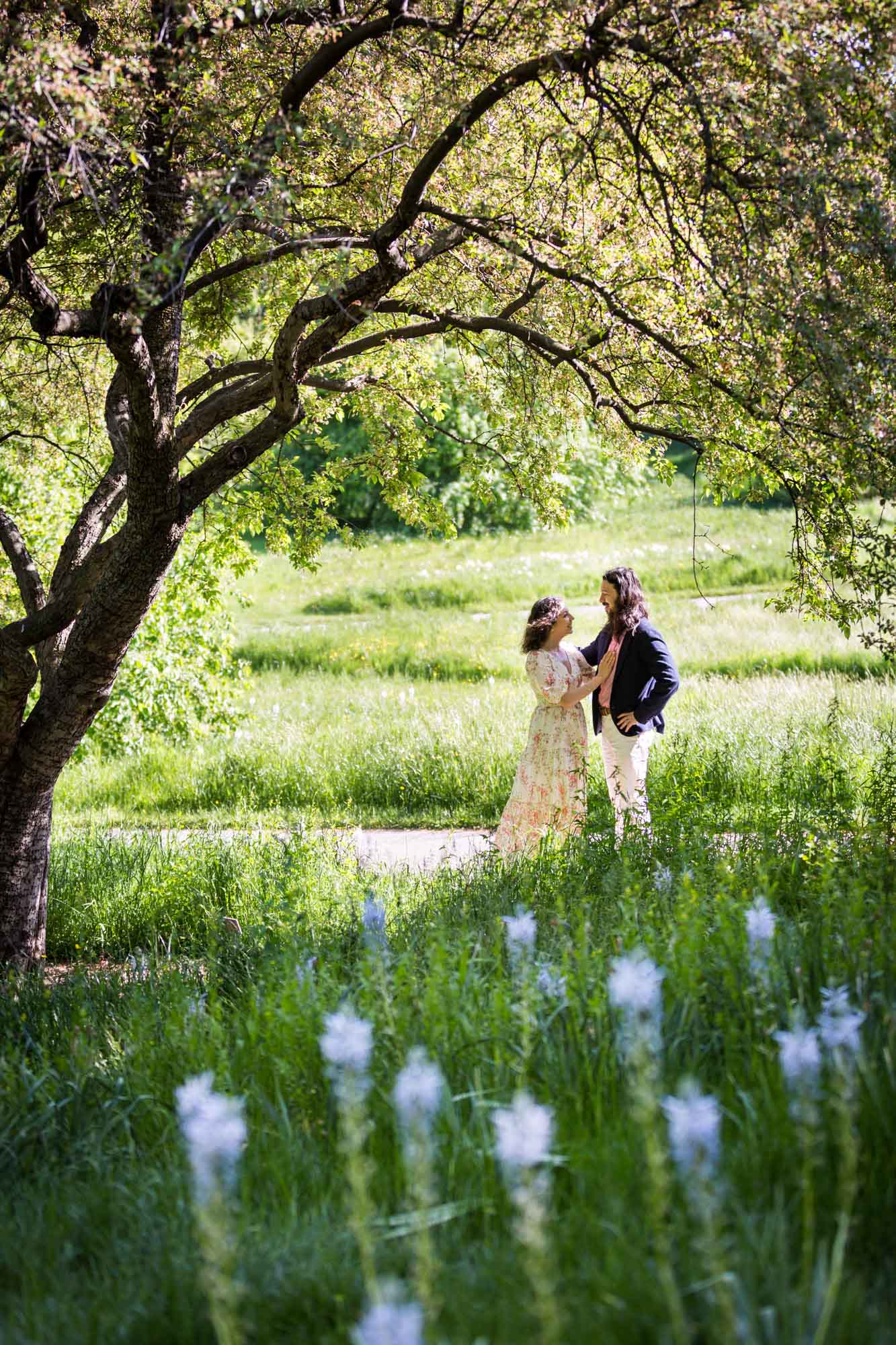Couple standing in pathway under tree behind blue flowers for an article on the best San Antonio Botanical Garden photo locations