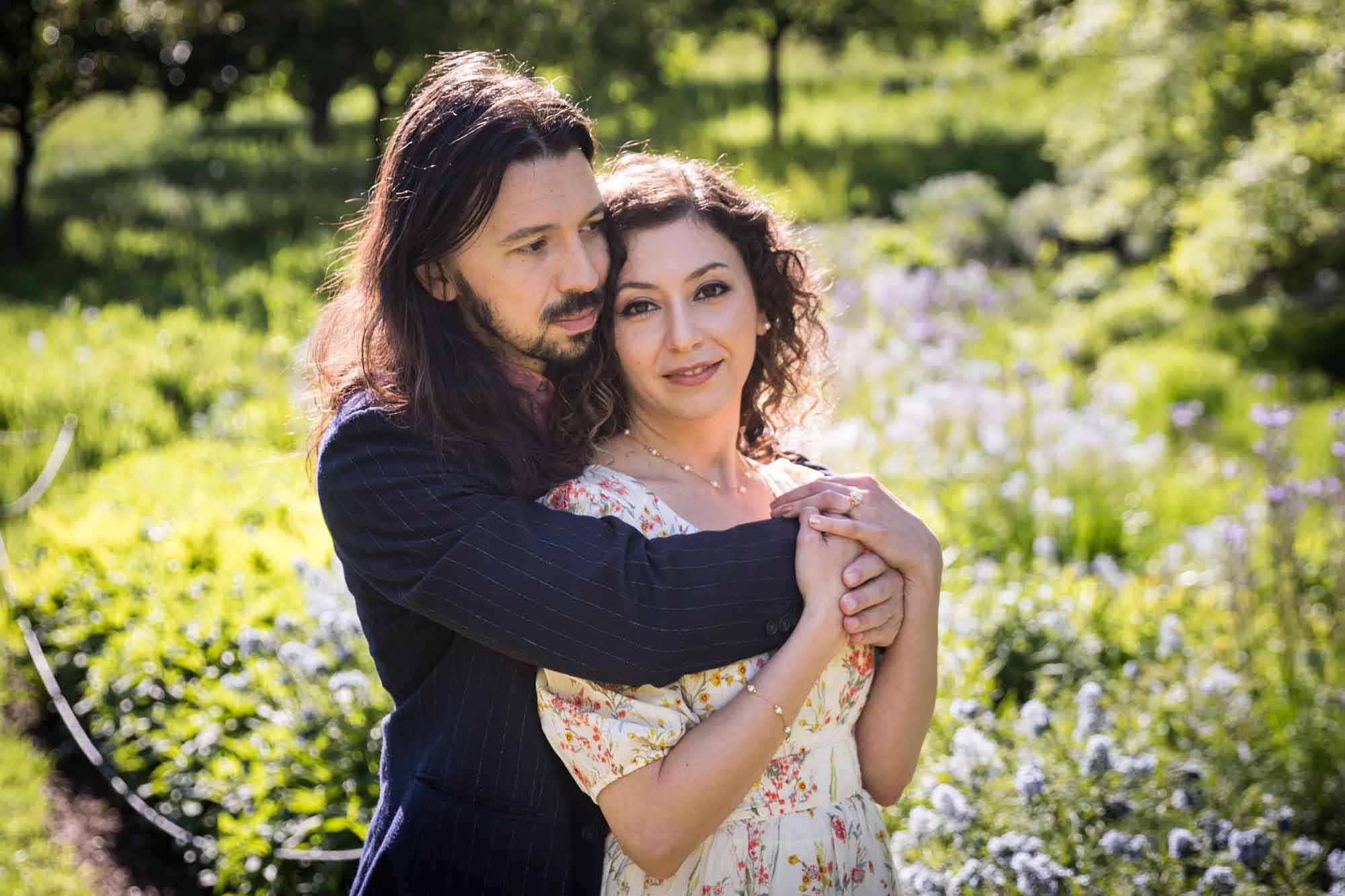 Man hugging woman from behind in front of purple and blue flowers and green bushes for an article on the best San Antonio Botanical Garden photo locations