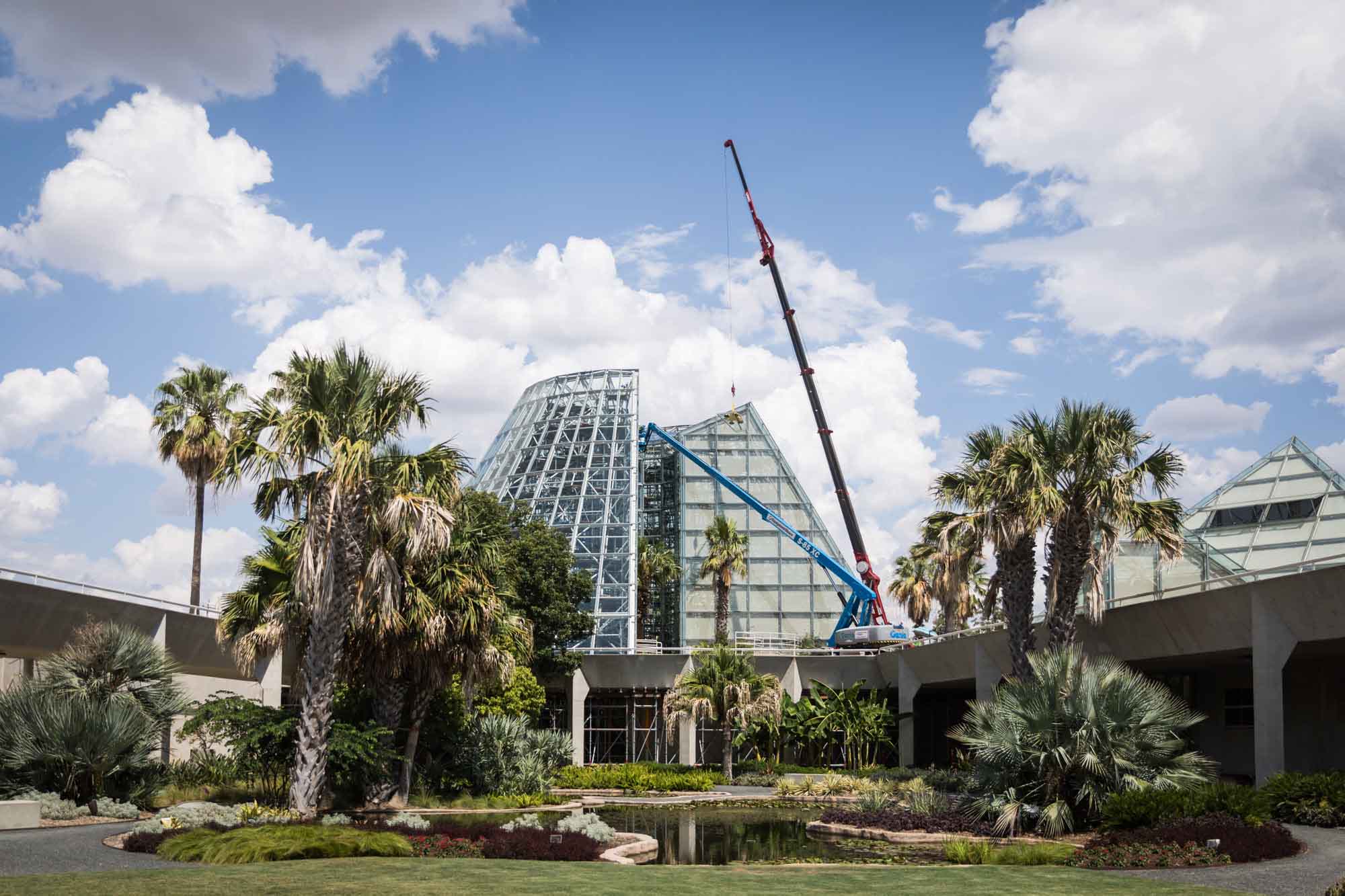 Wide view of exterior of Lucile Halsell Conservatory and lily pond with large construction crane working on interior and blue sky and clouds above for an article on the best San Antonio Botanical Garden photo locations