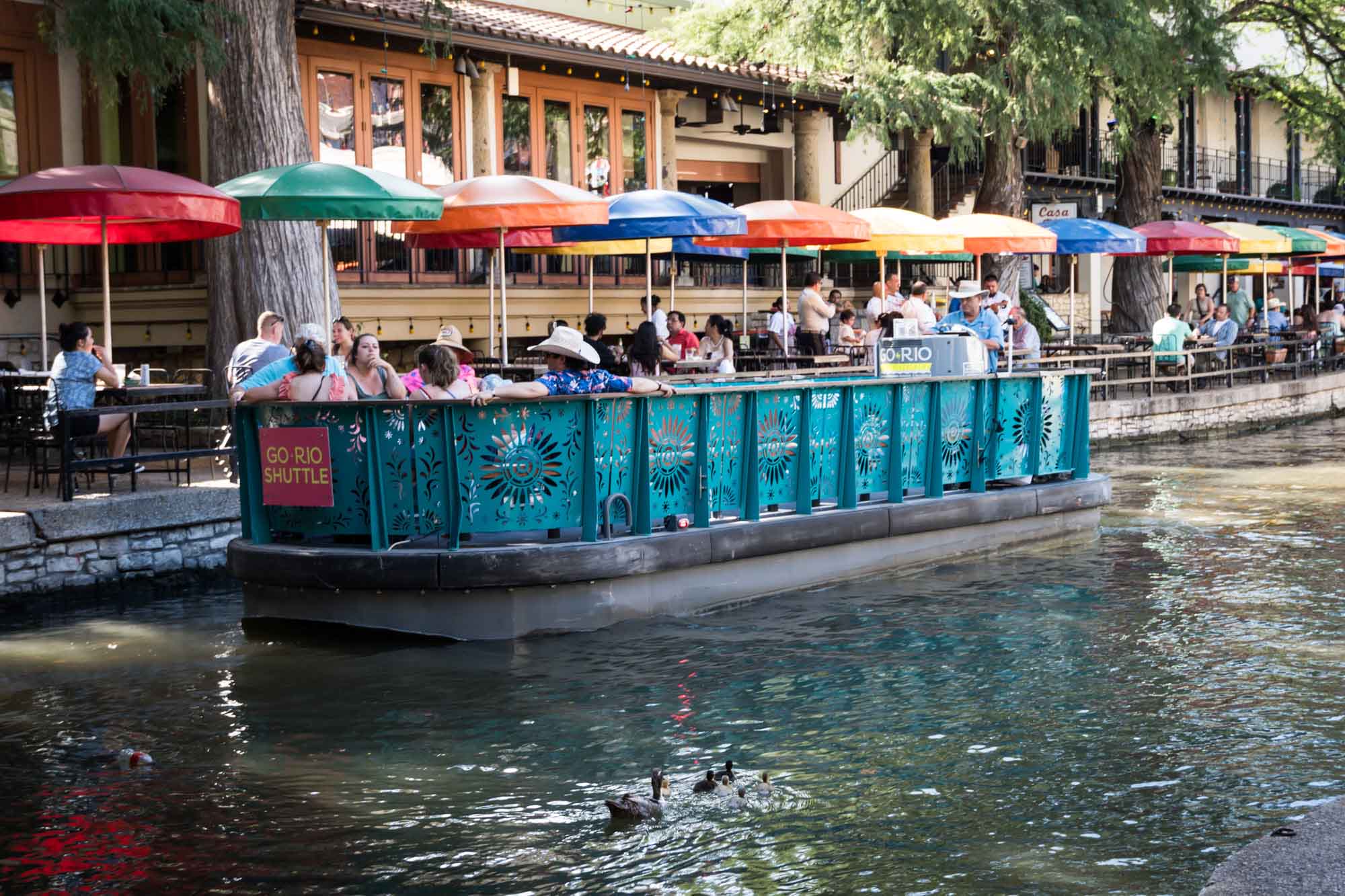 Aqua River Walk boat parked in front of colorful umbrellas of Casa Rio restaurant with mama duck and babies floating in front