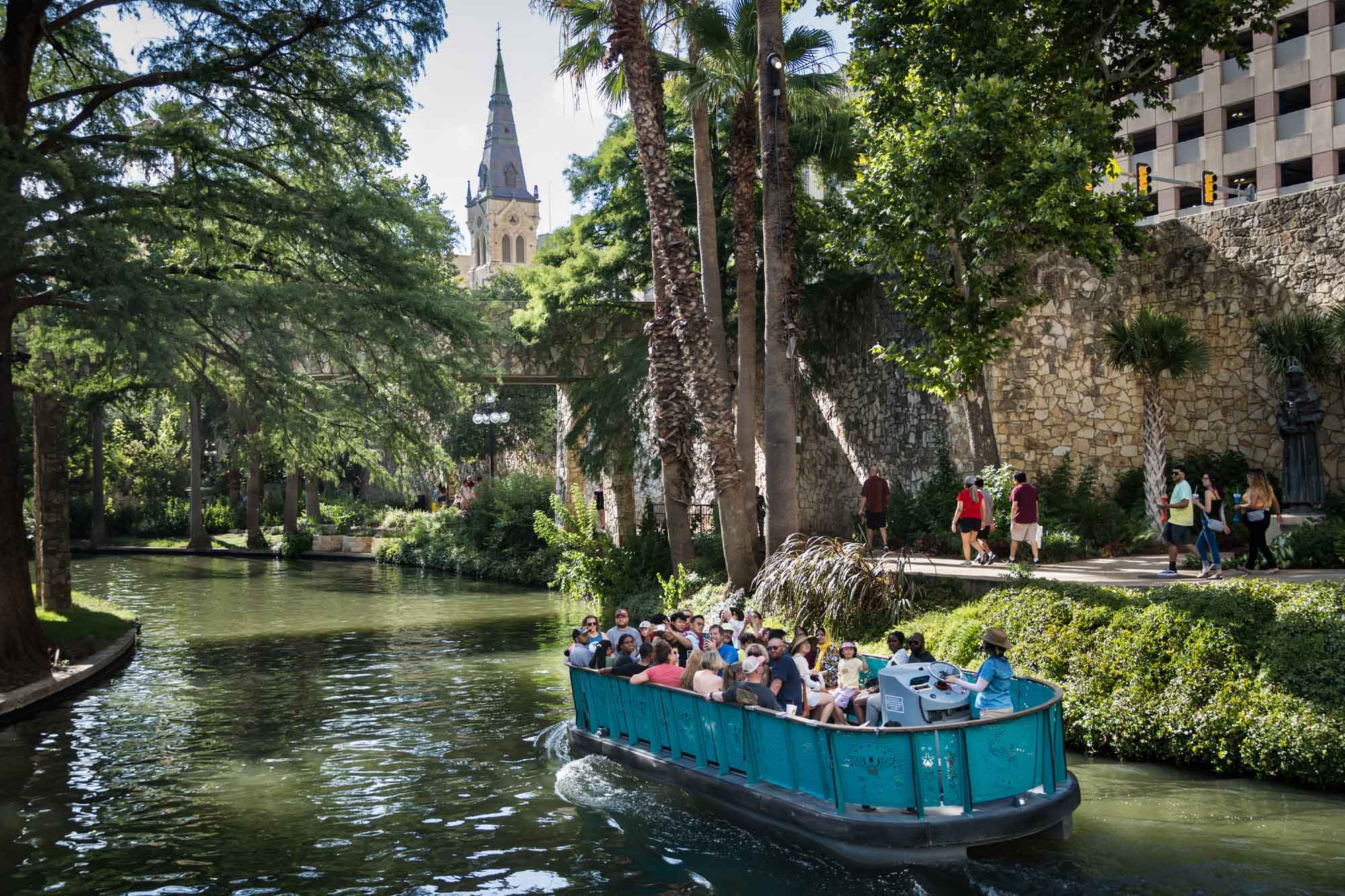 Aqua River Walk boat floating down the San Antonio River with stone bridge overhead and St. Joseph's Catholic Church in the distance