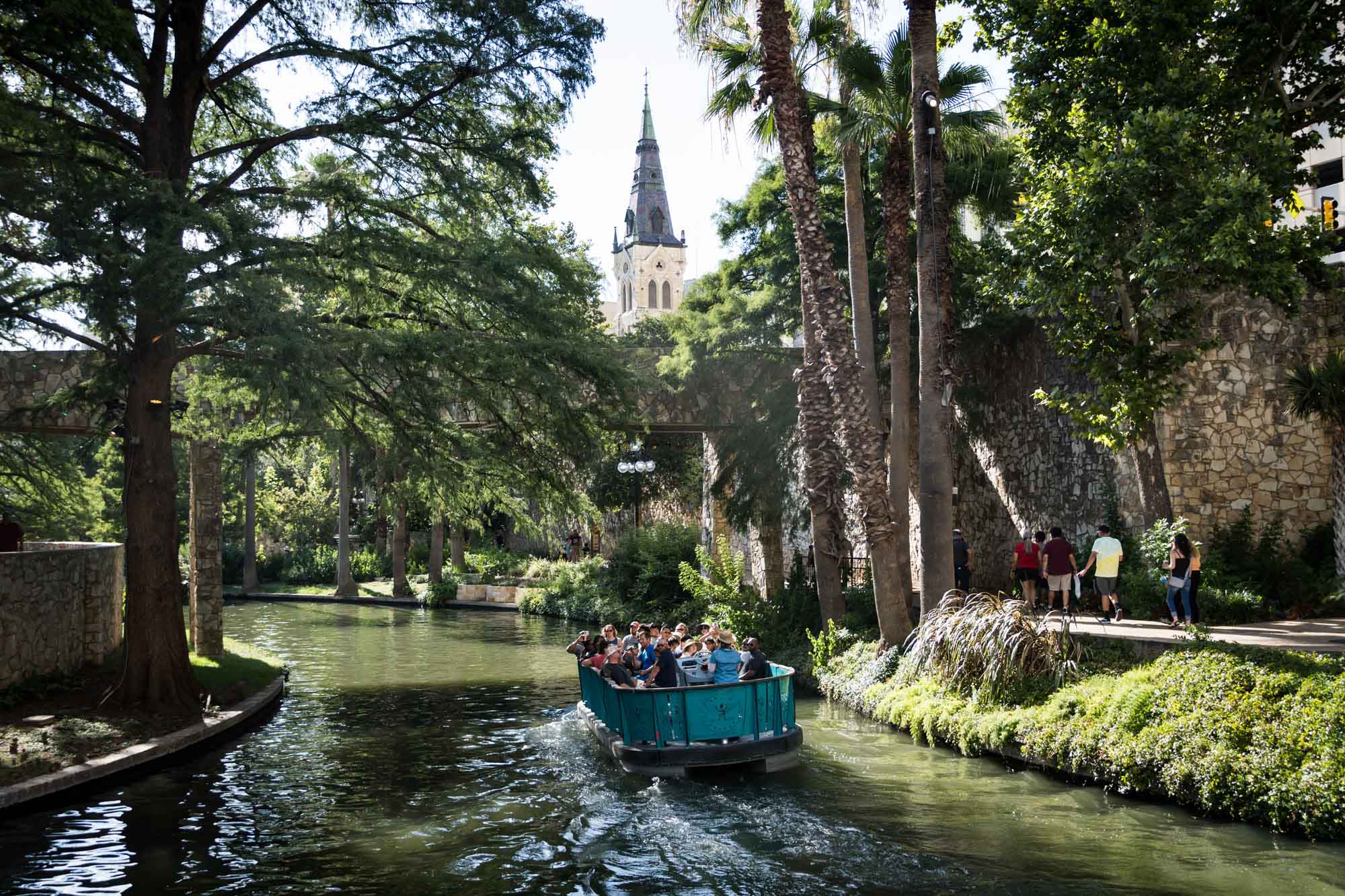 Aqua River Walk boat floating down the San Antonio River with stone bridge overhead and St. Joseph's Catholic Church in the distance