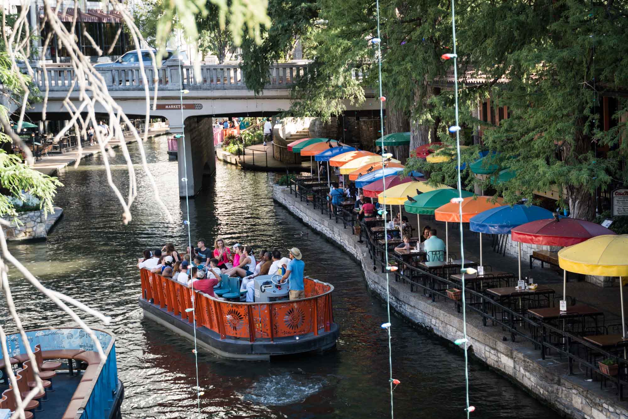 Orange River Walk boat full of tourists floating down the San Antonio River beside the colorful umbrellas of the Casa Rio restaurant