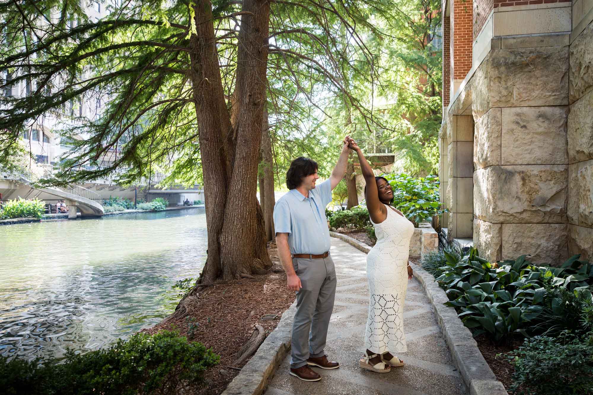 African American woman wearing white dress dancing with white fiance on River Walk pathway beside river for an article on how to get married on a River Walk boat