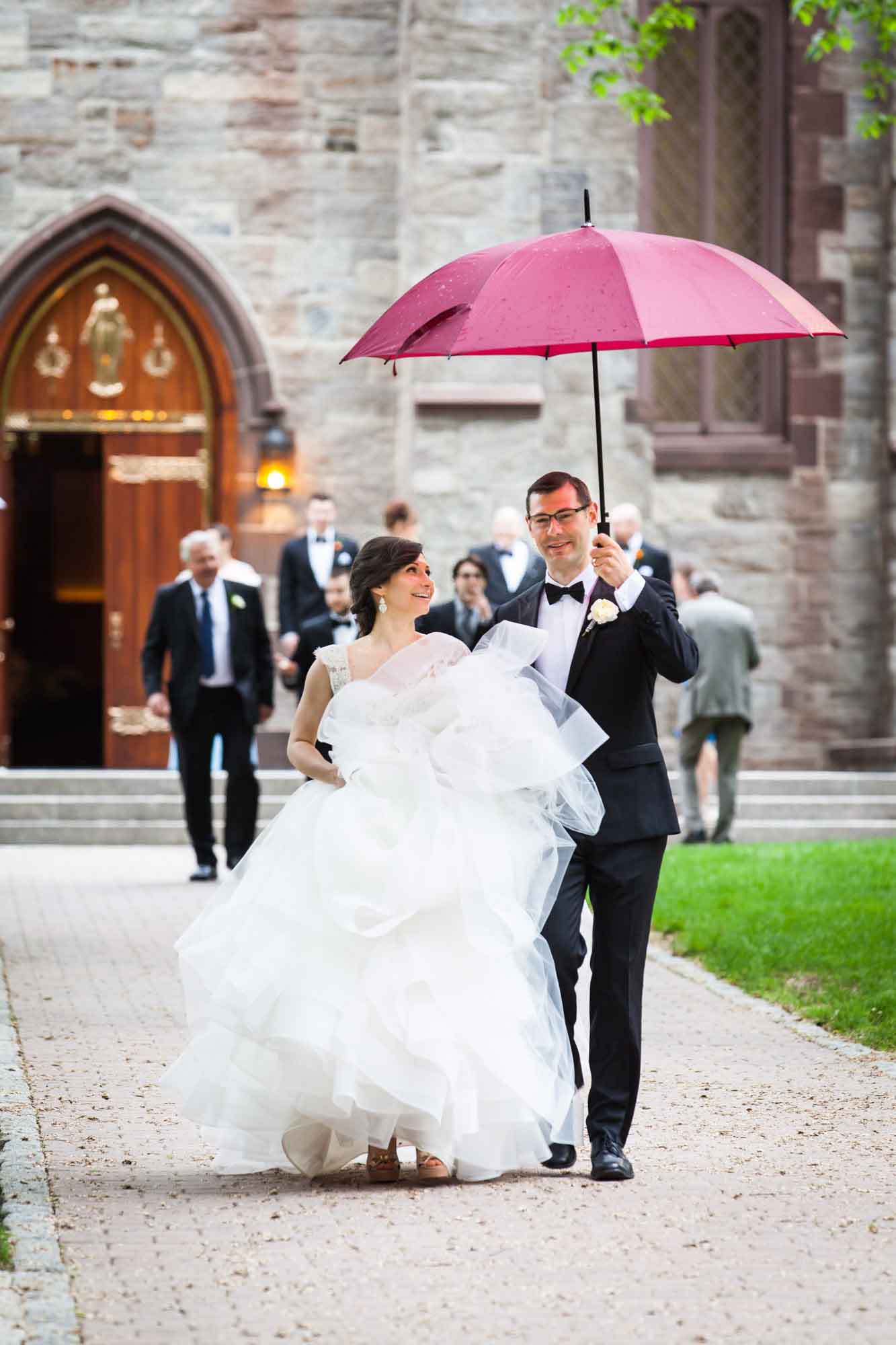Bride and groom leaving a Fordham University Church wedding ceremony under red umbrella for an article on what to have ready when the wedding photographer arrives