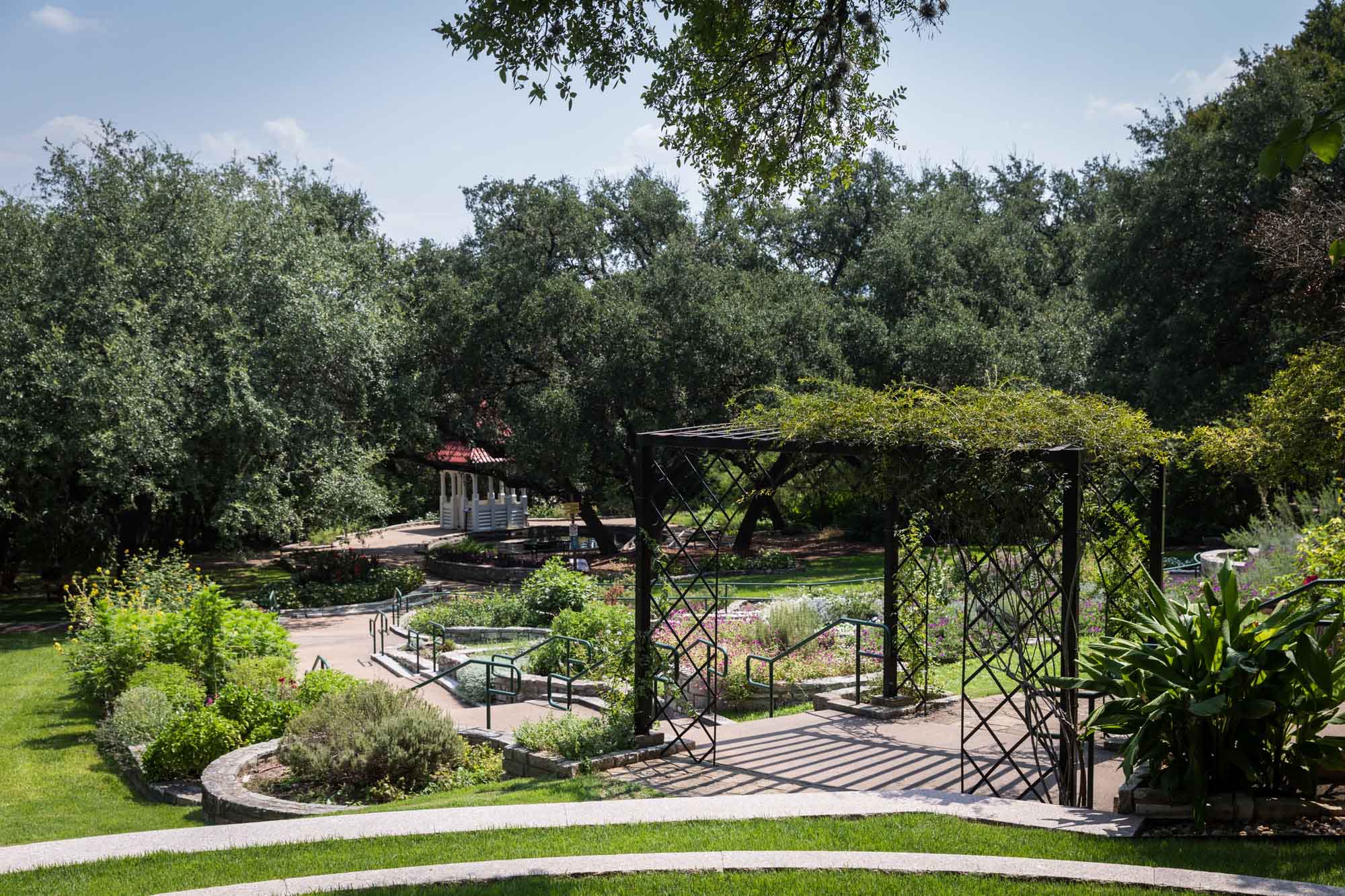 Black lattice-work gazebo covered in vines with gazebo in background and flower beds for an article on the best Zilker Botanical Garden photo locations
