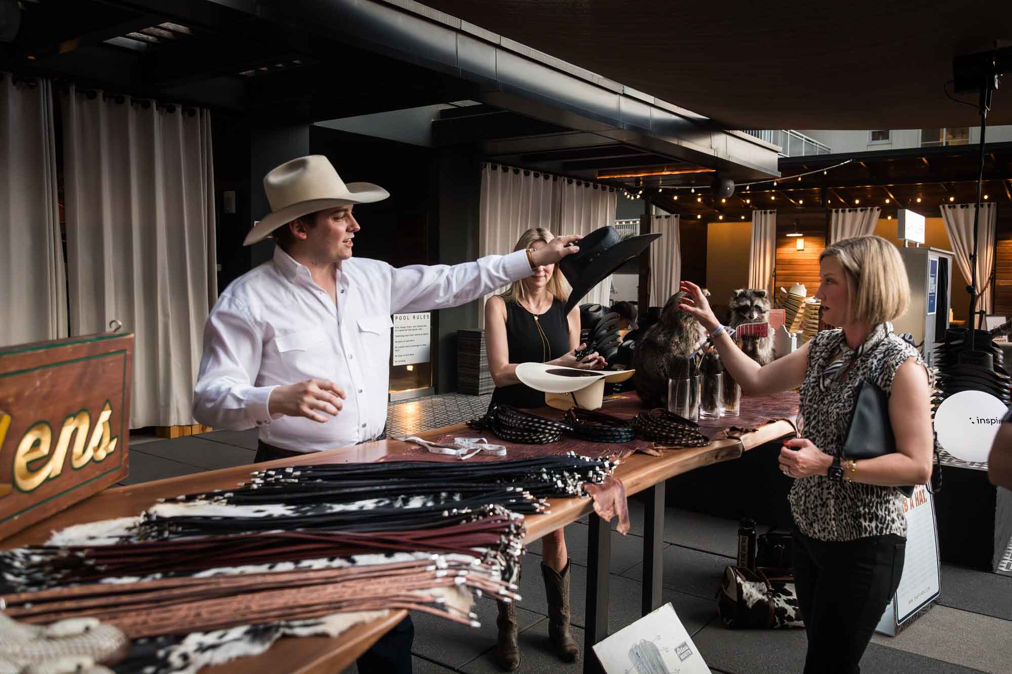 Man in white shirt wearing cowboy hat handing a black cowboy hat to a woman across a table of belts during a Hotel Van Zandt corporate event
