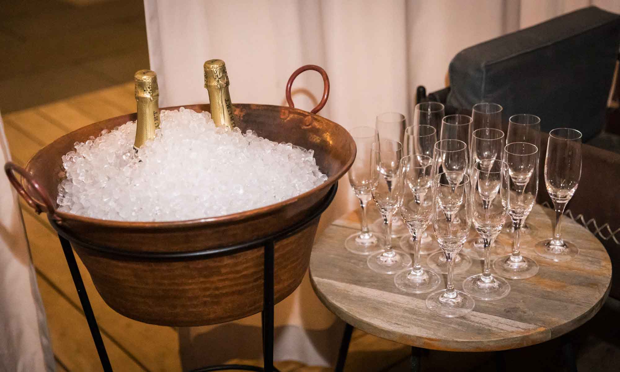 Large container of ice with two champagne bottles sticking out next to table with several champagne glasses