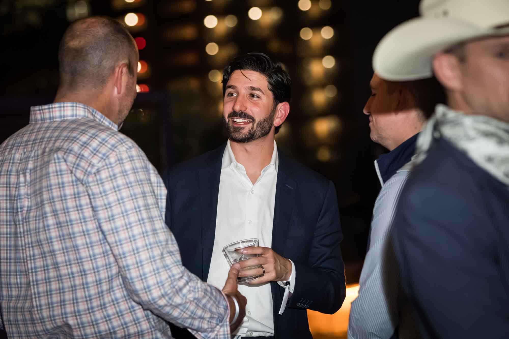 Smiling man with black hair wearing blue blazer and white shirt holding drink talking to man wearing plaid shirt during a Hotel Van Zandt corporate event