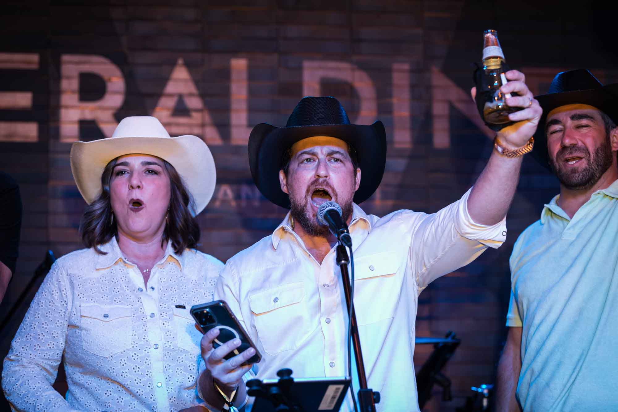 Man wearing black cowboy hat holding cell phone and beer bottle in the air talking into microphone with man and woman behind him during a Hotel Van Zandt corporate event
