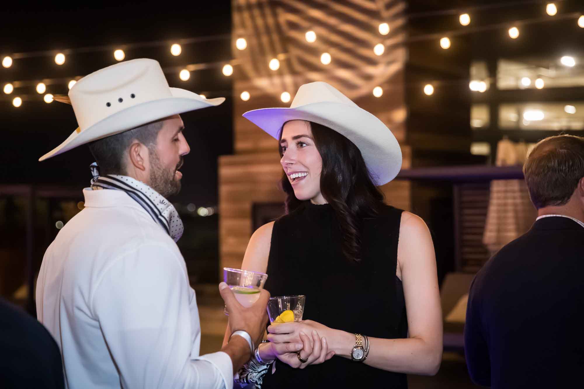 Couple wearing white cowboy hats talking in front of string lights holding drinks during a Hotel Van Zandt corporate event